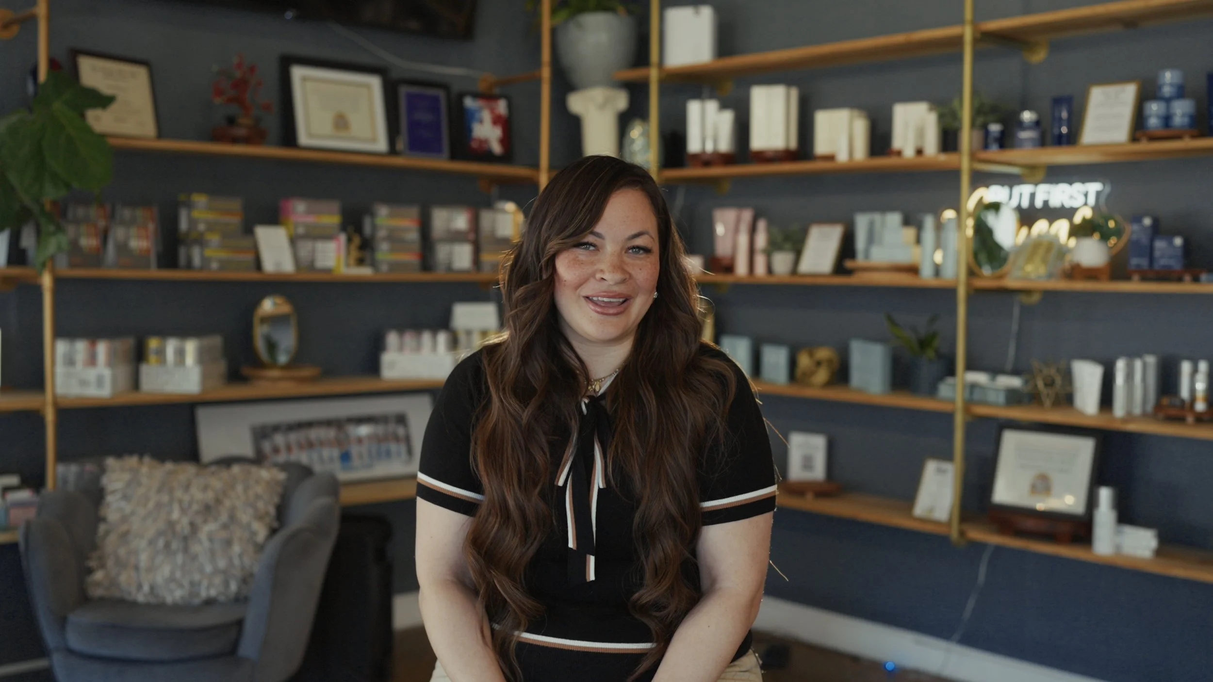 A woman with long wavy brown hair, wearing a black shirt with a bow, is sitting in a modern, well-decorated home or office space with bookshelves, framed pictures, and a neon sign in the background.