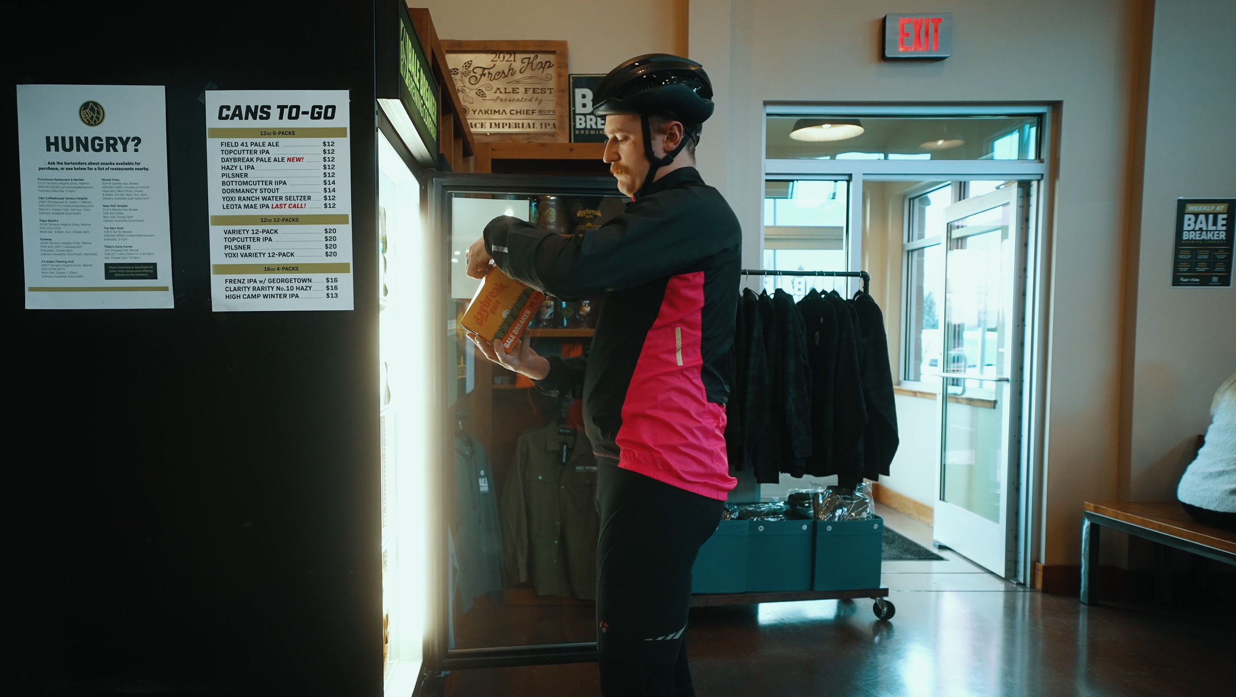 A man wearing a black helmet and black and red athletic clothing is standing inside a store, looking at a box of beer. The store has a menu on the wall listing various craft beers and a doorway leading outside.