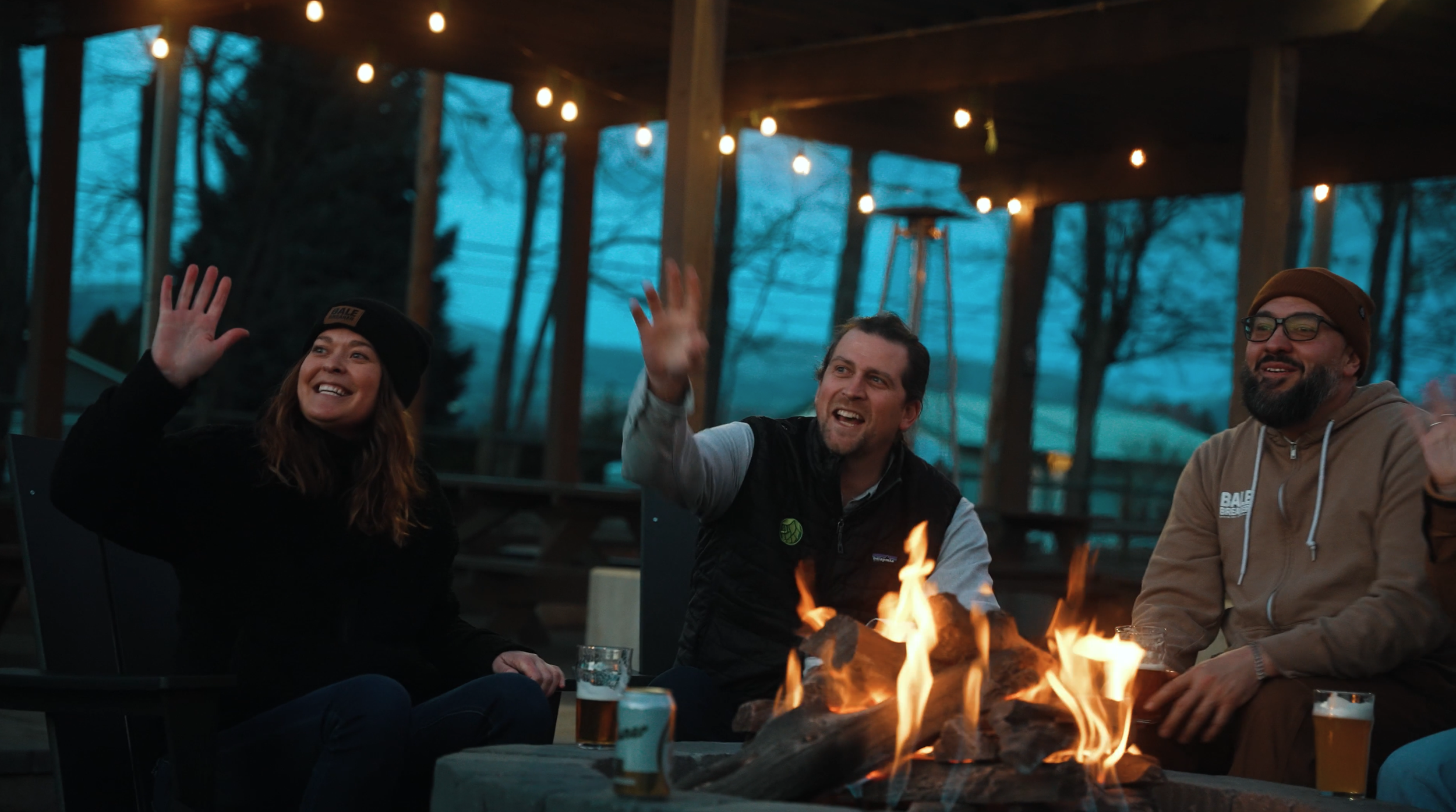 Three people sitting around a fire pit outdoors during dusk, smiling and waving. String lights hang above them, and trees can be seen in the background.