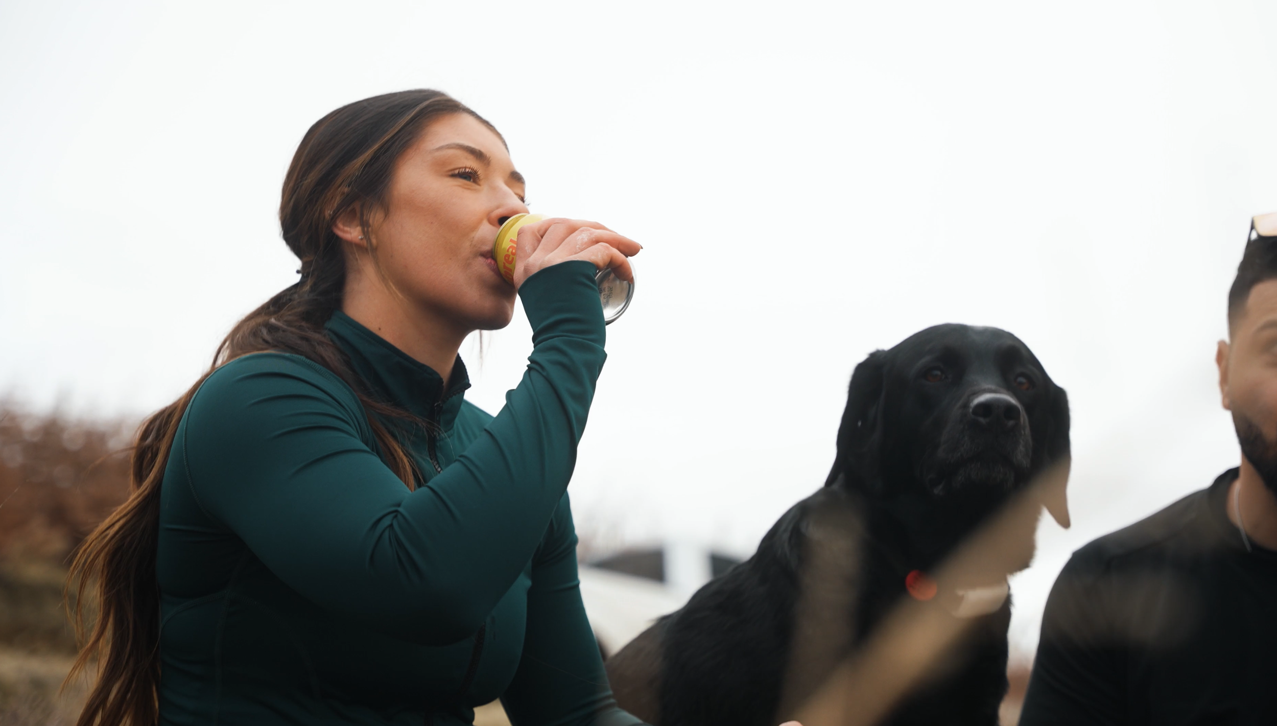 A woman in a teal jacket drinks from a can while outdoors, with a black and tan dog and a person with sunglasses nearby.