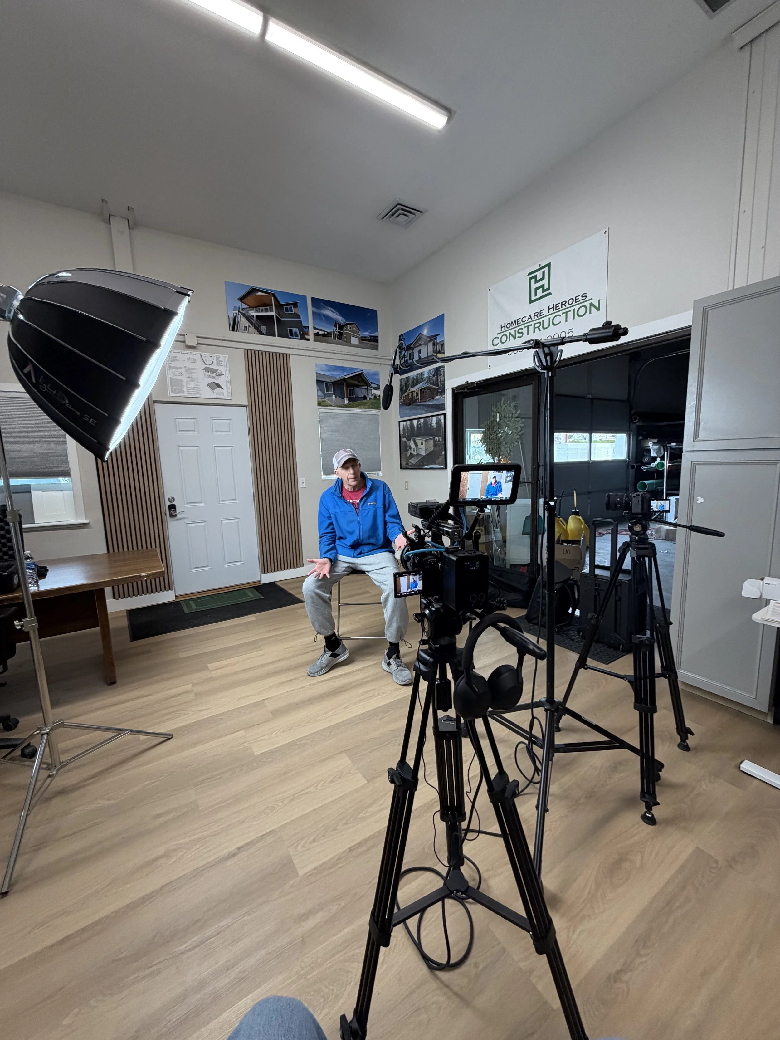 A man sitting on a chair in a room, being filmed with professional video equipment, including a camera on a tripod, softbox lights, and microphones, with display images and a 'Homecare Heroes Construction' sign on the wall behind him.