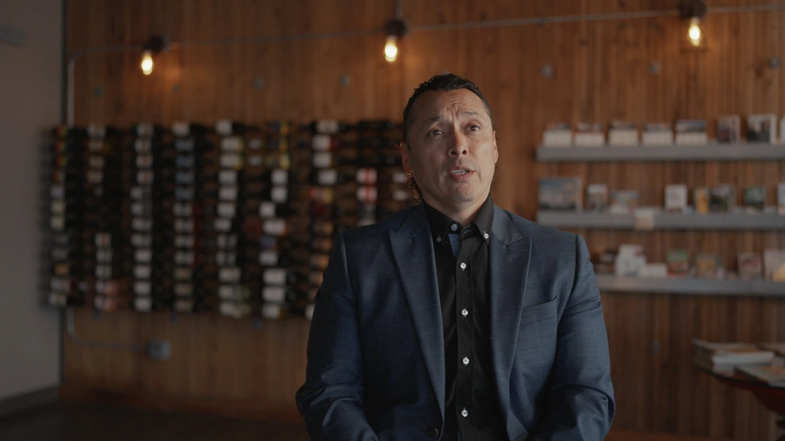 A man in a dark suit and black shirt sitting in front of a wooden wall with shelves holding books or magazines.