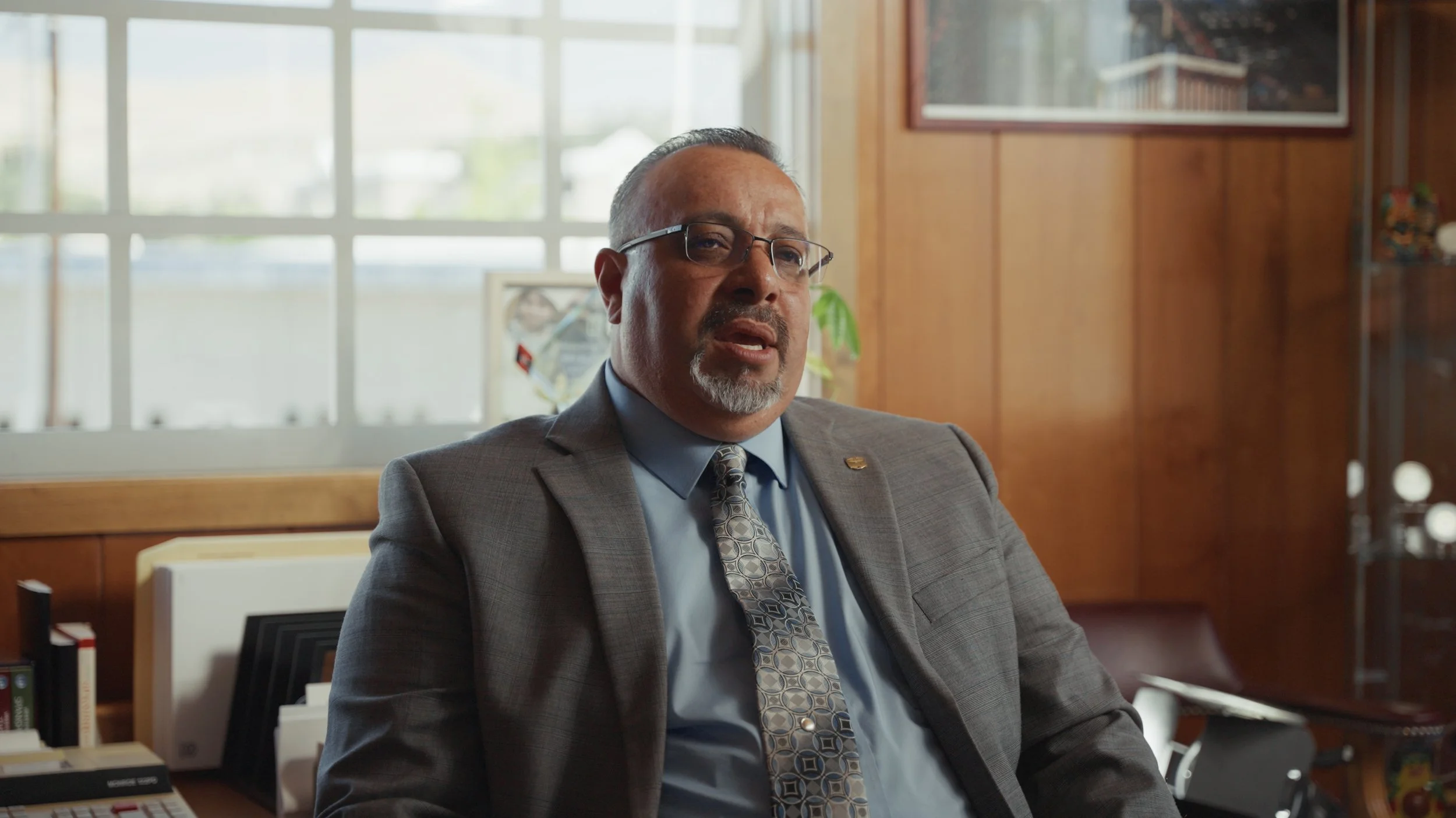 A man in a suit and glasses sitting in an office, talking, with a window and wood-paneled walls in the background.