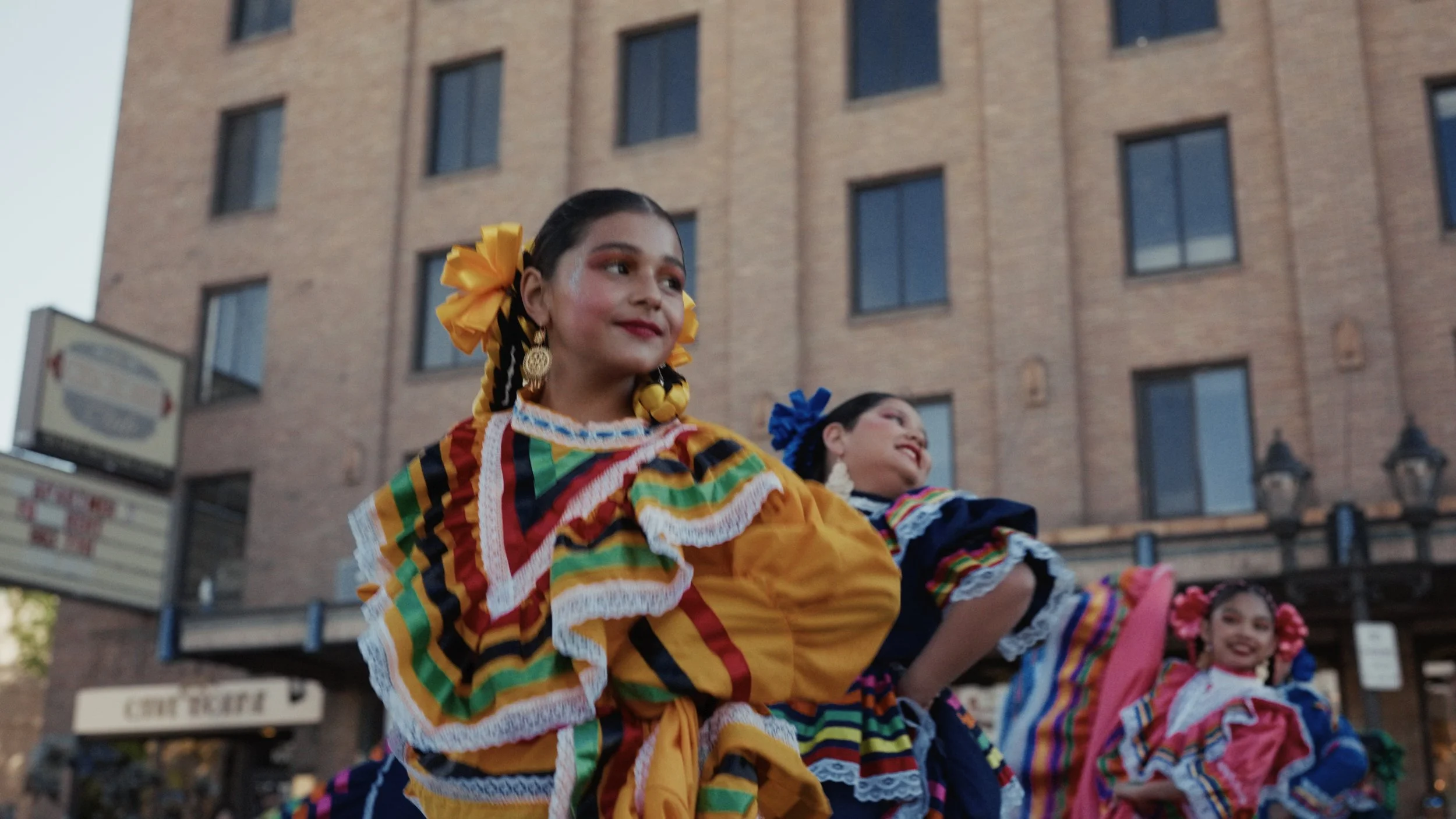 Women dressed in traditional Mexican colorful folkloric dresses performing a dance outdoors in front of a brick building during a cultural event.