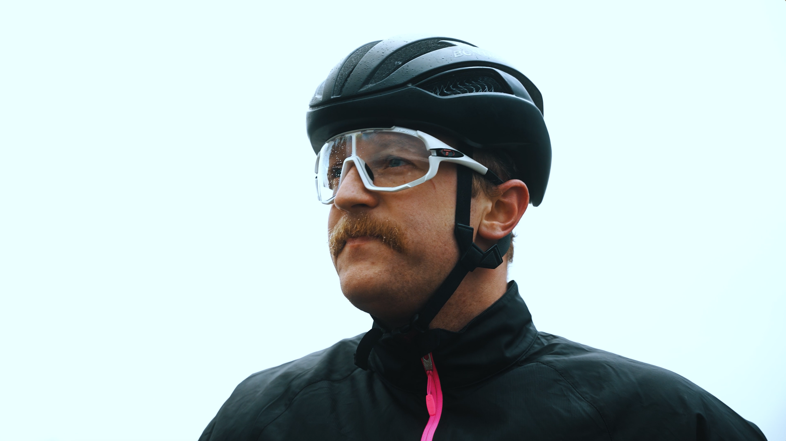 A man wearing a black bicycle helmet and sports glasses, looking to the side, against a light sky background.