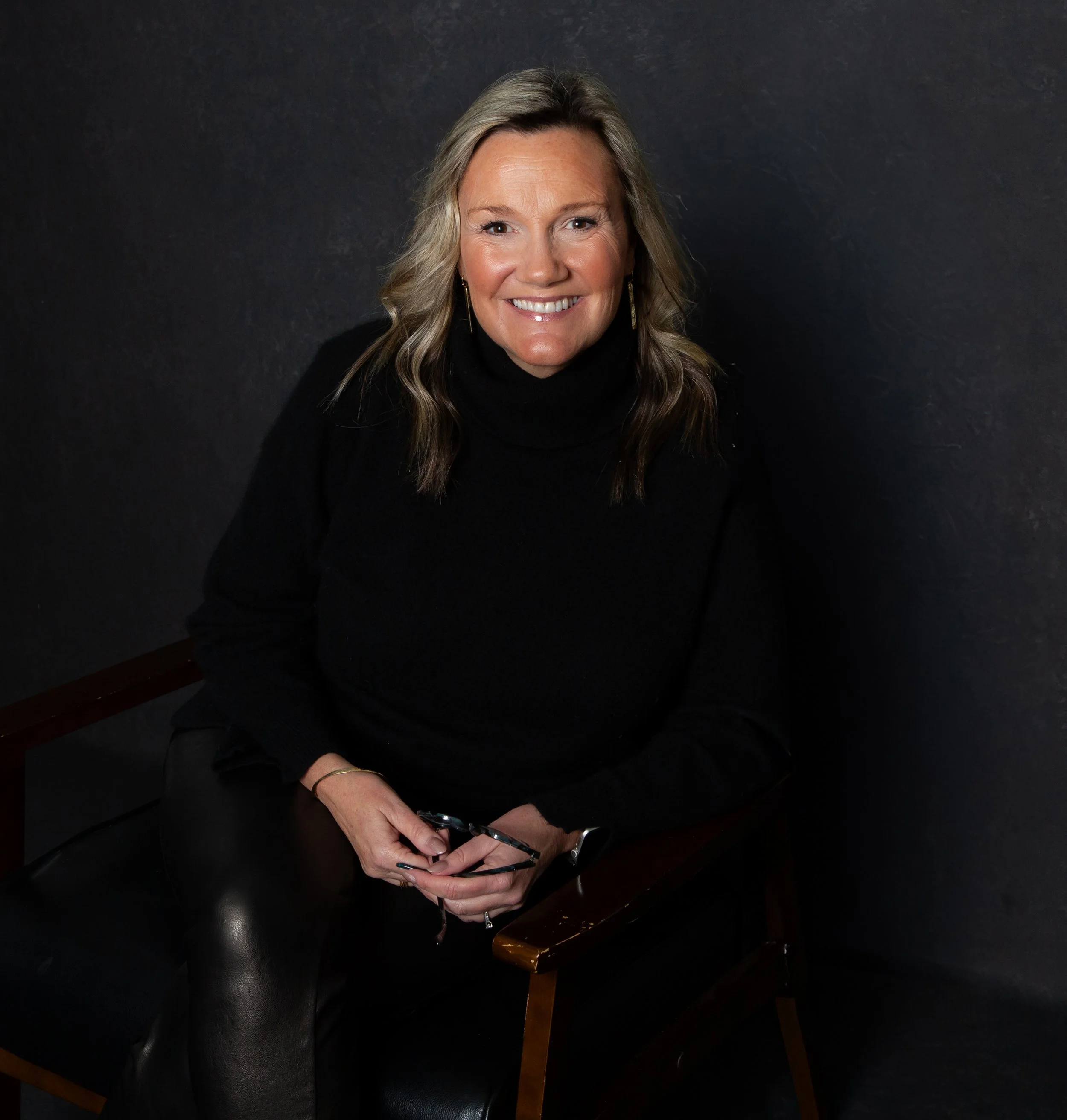 Erin Herringshaw wearing a black turtleneck sweater, sitting on a wooden chair against a dark background, smiling and holding eyeglasses.