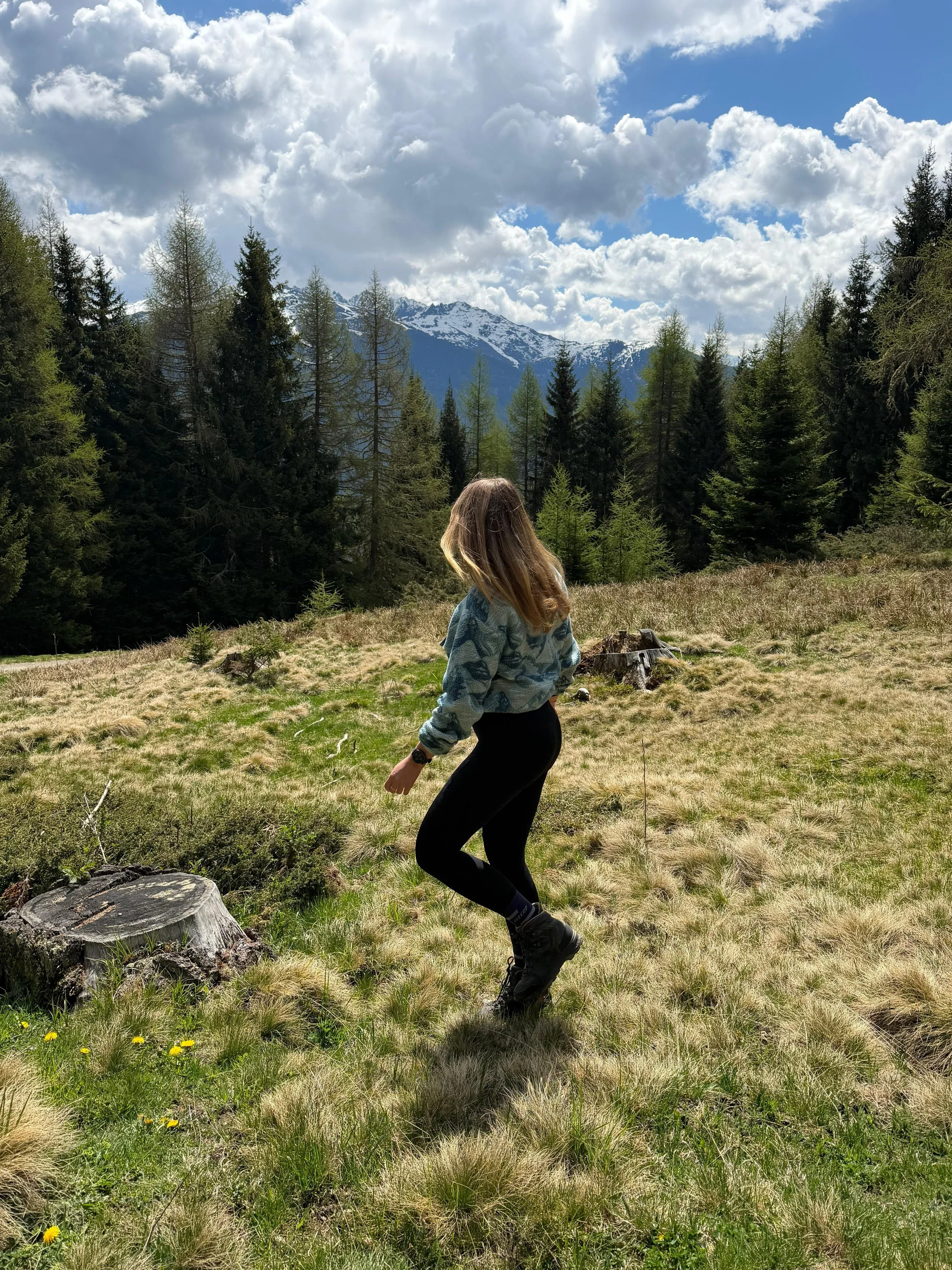 Eine Frau in Bewegung auf einer Wiese mit Bäumen vor Bergen und Wolken am Himmel.