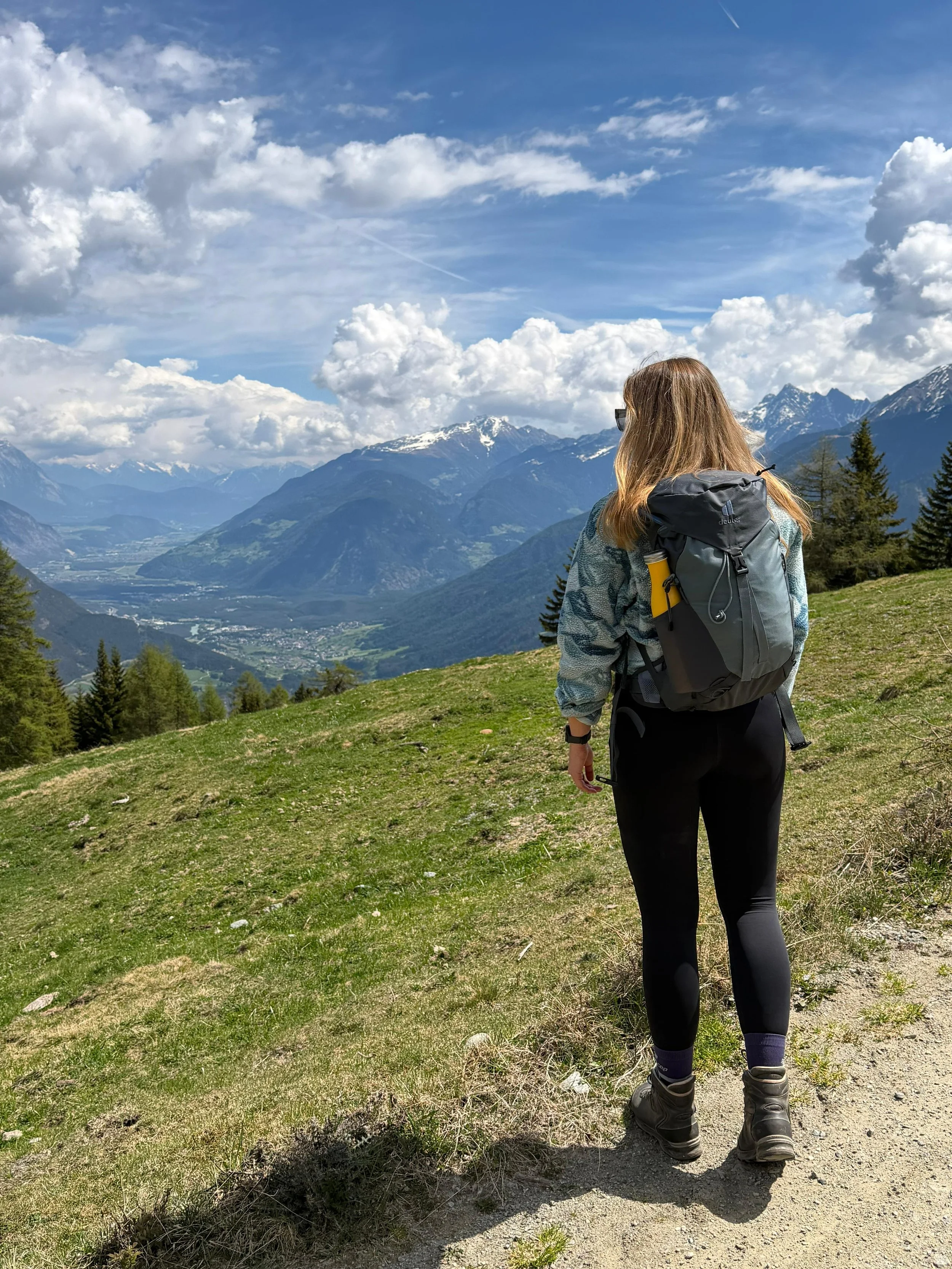 Eine Frau mit langem, braunem Haar, Sonnenbrille, grauem Rucksack und Wanderschuhen steht auf einem Weg in einer bergigen Landschaft, umgeben von grünen Wiesen, Tannenbäumen und schneebedeckten Bergen im Hintergrund, bei schönem Wetter mit vielen Wolken am Himmel.