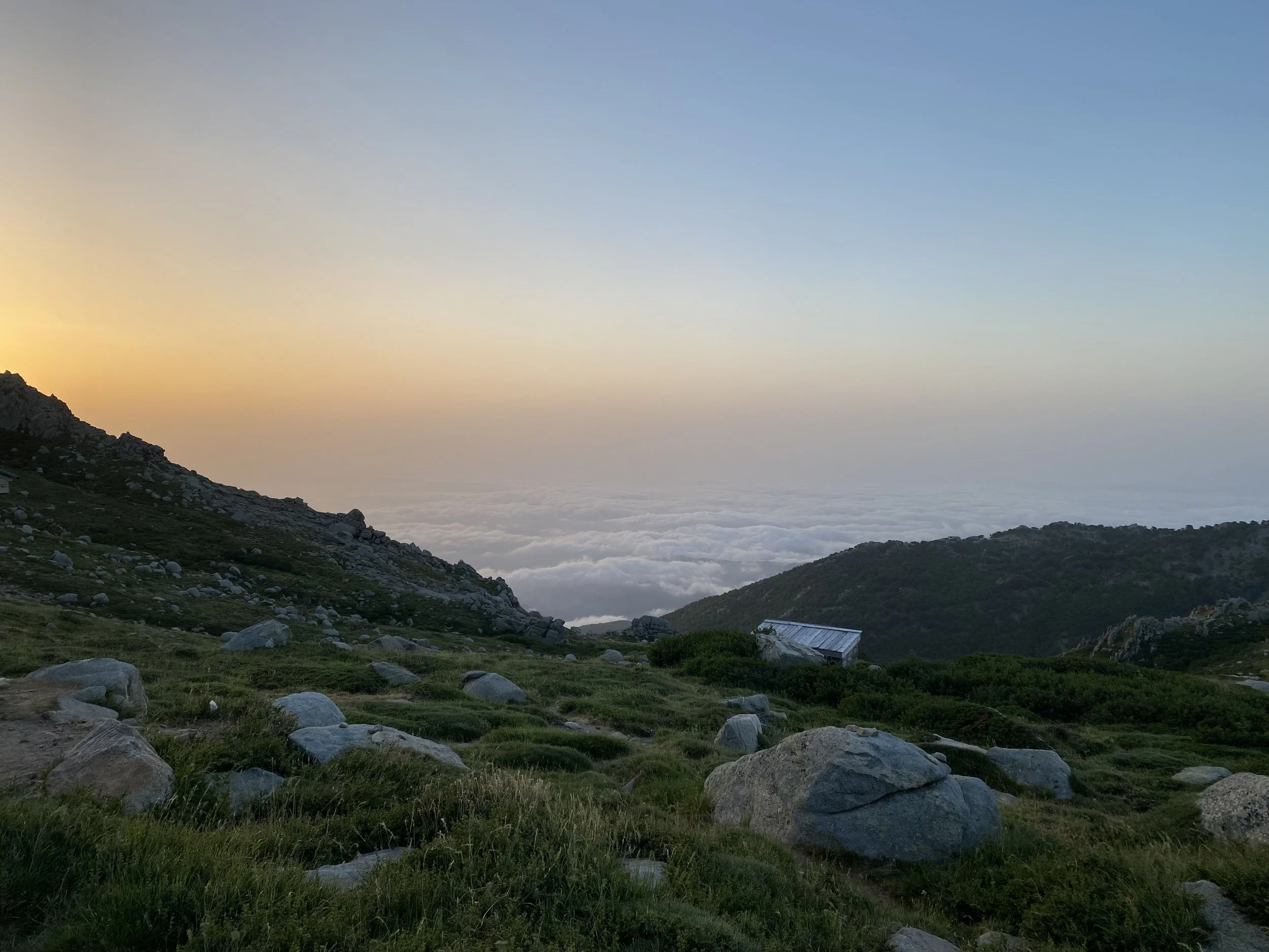 Landschaft mit bewaldeter Hügellandschaft, Wolkendecke im Tal, sanftem Sonnenlicht bei Sonnenuntergang oder Sonnenaufgang, einige Häuser im Vordergrund.