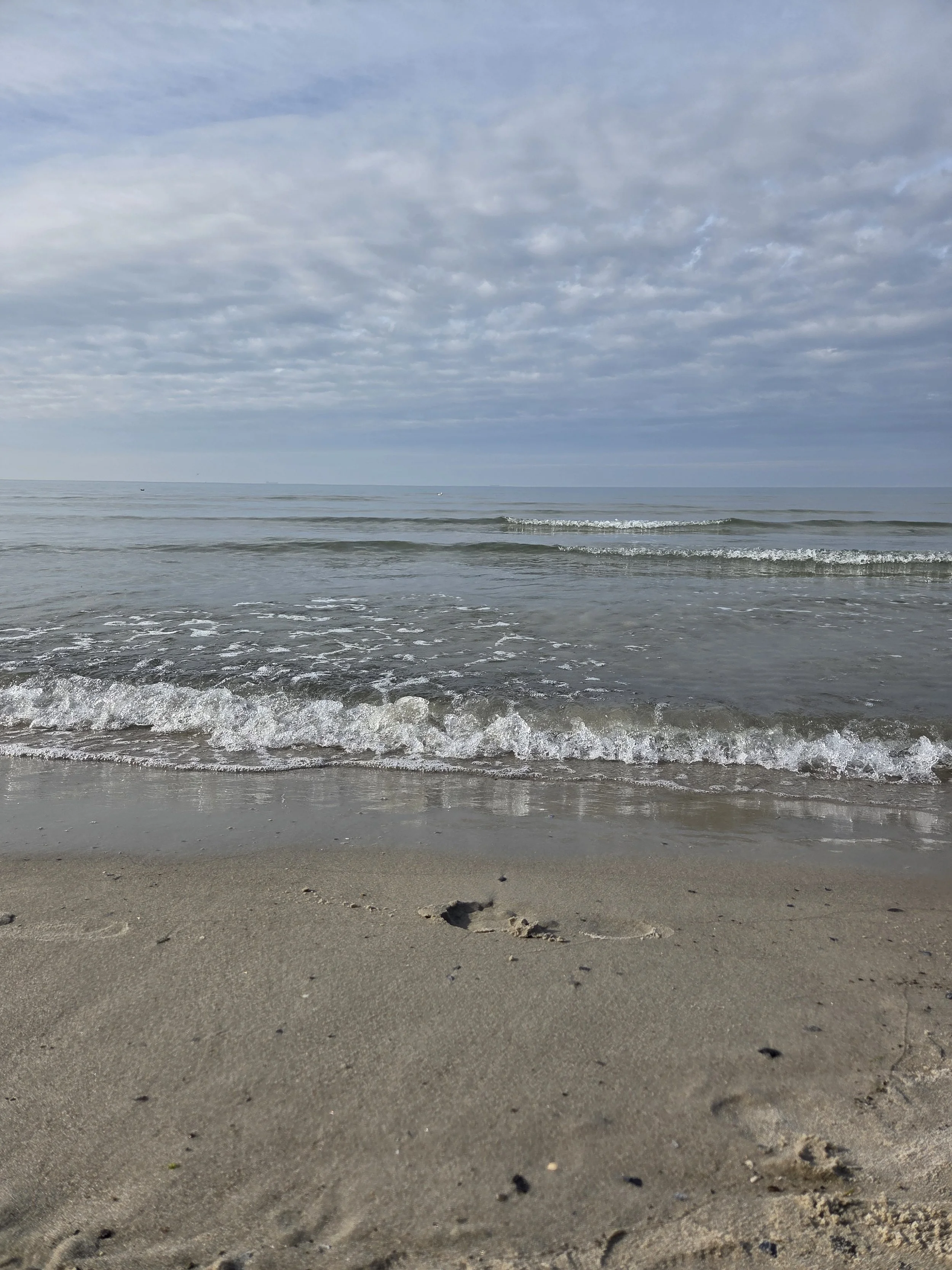 Strand mit Sand, kleinen Steinchen und Fußabdruck, sanfte Wellen im Meer, bewölkter Himmel.
