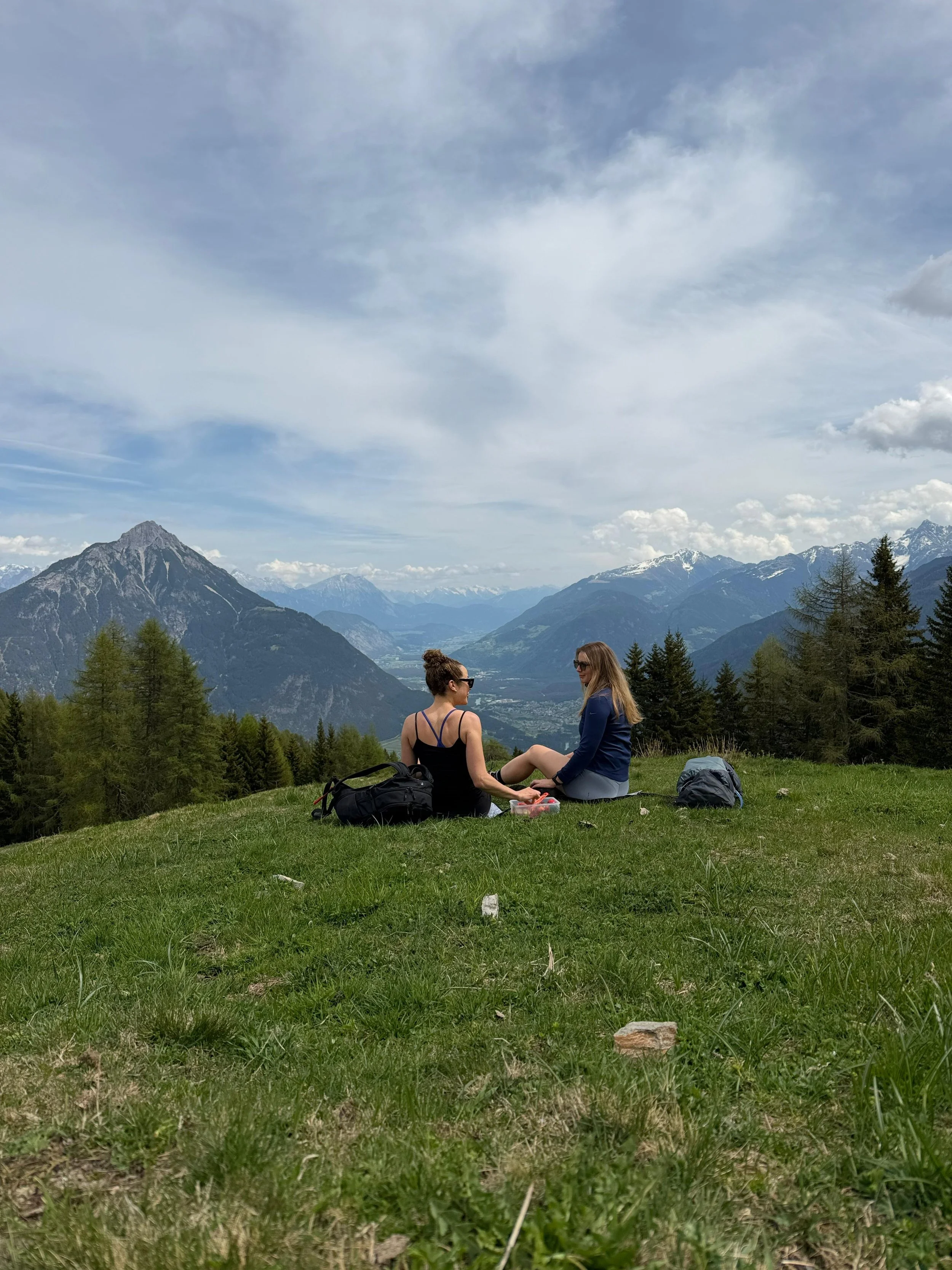 Zwei Frauen sitzen auf einer grünen Wiese in den Bergen, umgeben von Tannenbäumen und hohen Bergen im Hintergrund, während sie miteinander sprechen.