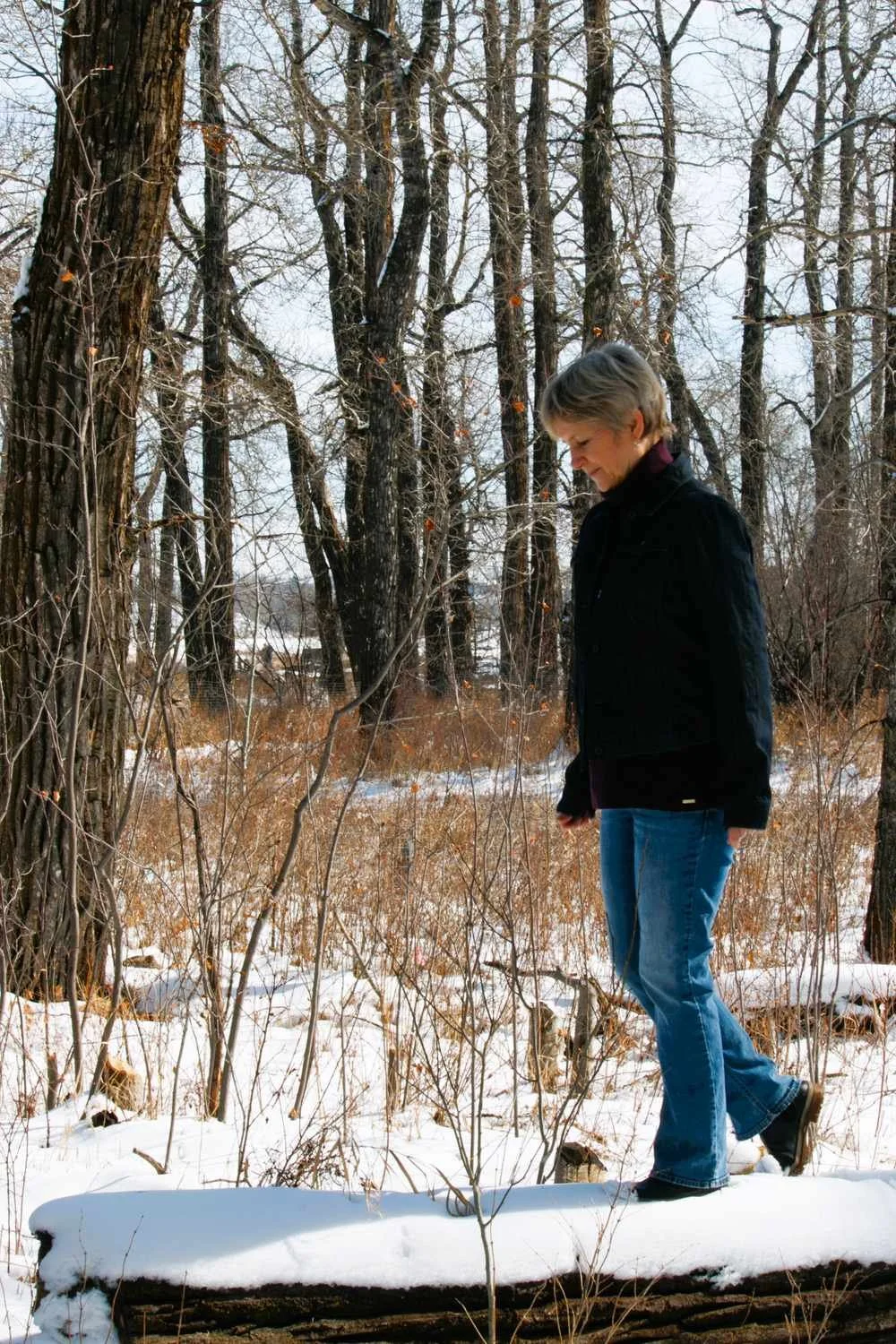 Liz Rees walking along a snow-dusted log through a bare winter forest, an image of the contemplative journey through the seasons of the soul