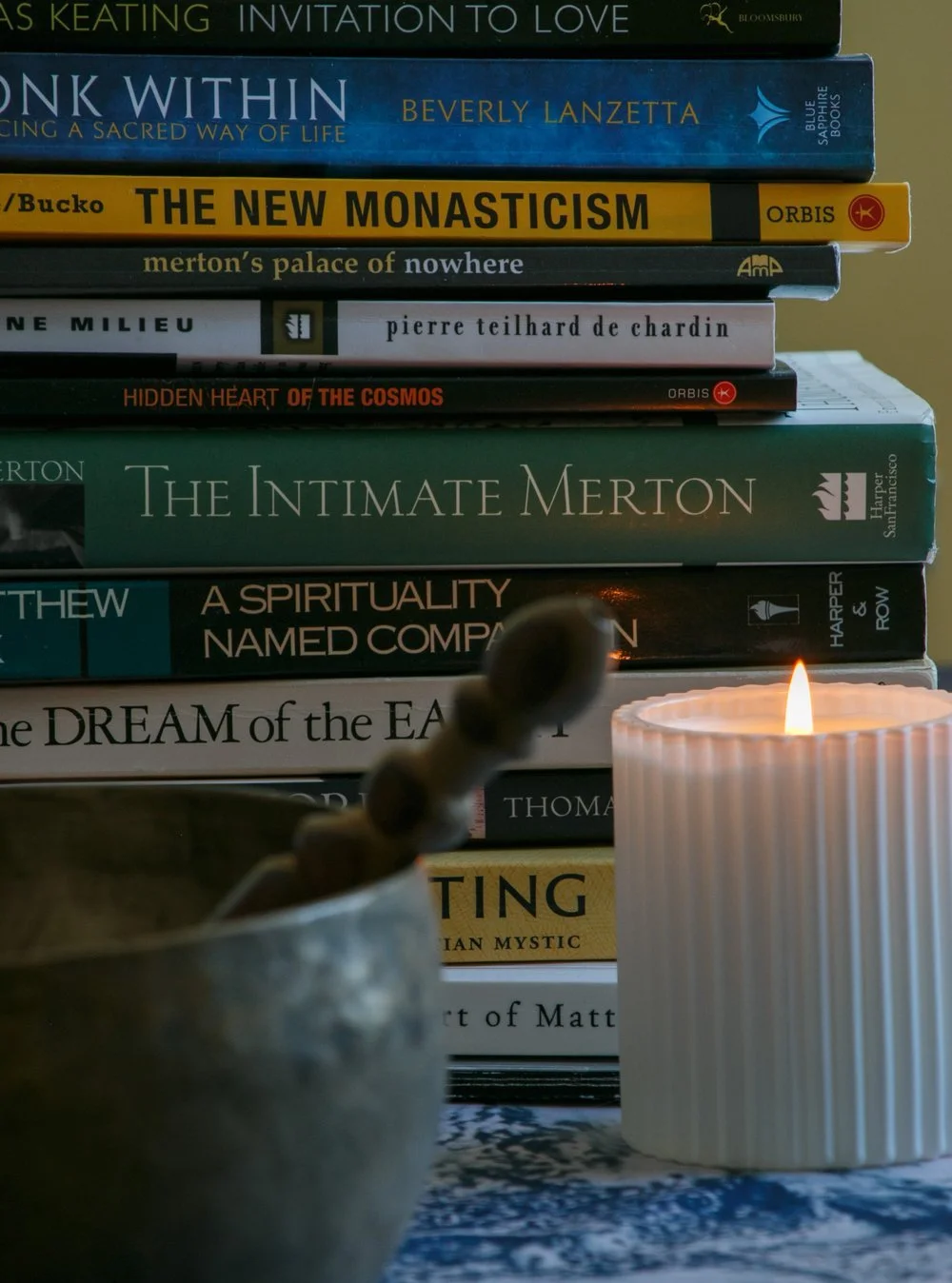 Stack of contemplative books by Thomas Merton, Beverly Lanzetta, Pierre Teilhard de Chardin and others, with a lit candle and singing bowl — grounding Liz's offerings in the Christian contemplative tradition