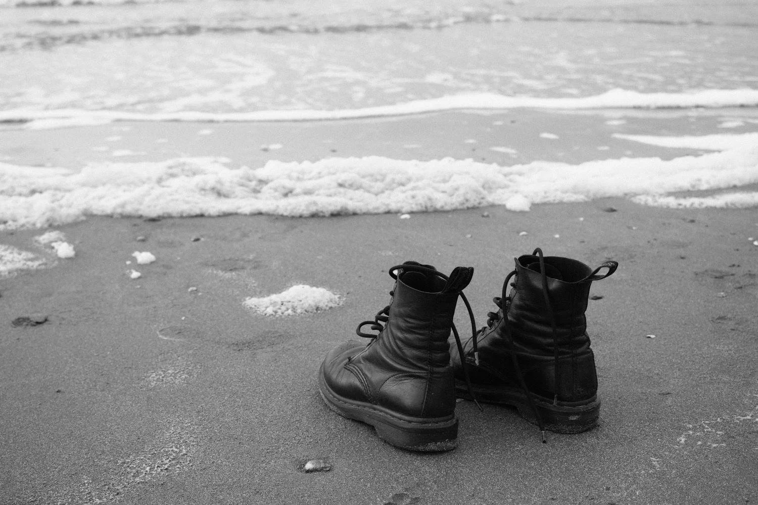 Black and white image of leather boots resting on wet sand at the ocean's edge, evoking the pilgrim soul's contemplative journey