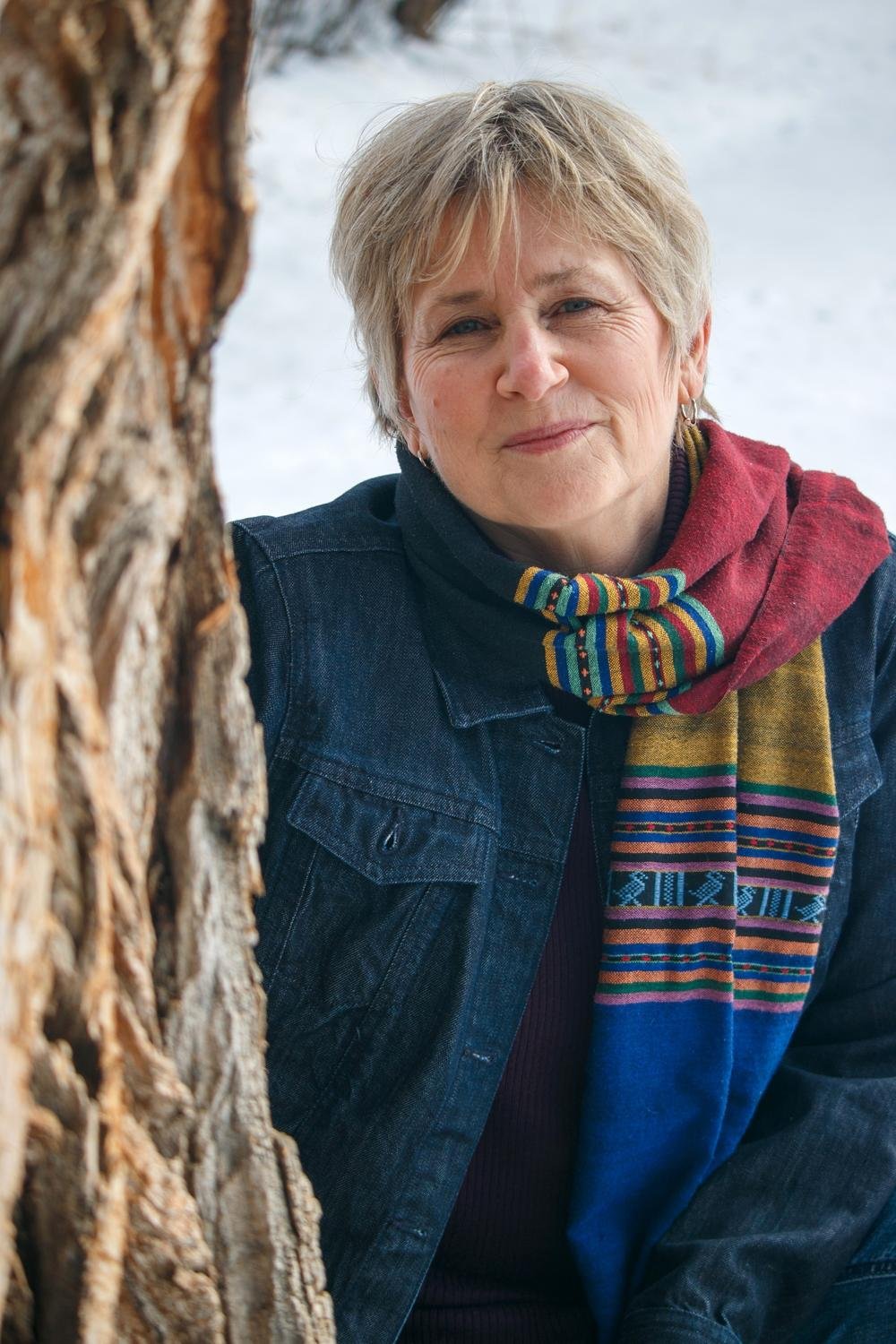 Portrait of spiritual director Liz Rees in a denim jacket and colourful striped scarf, leaning against a tree trunk on a snowy winter day