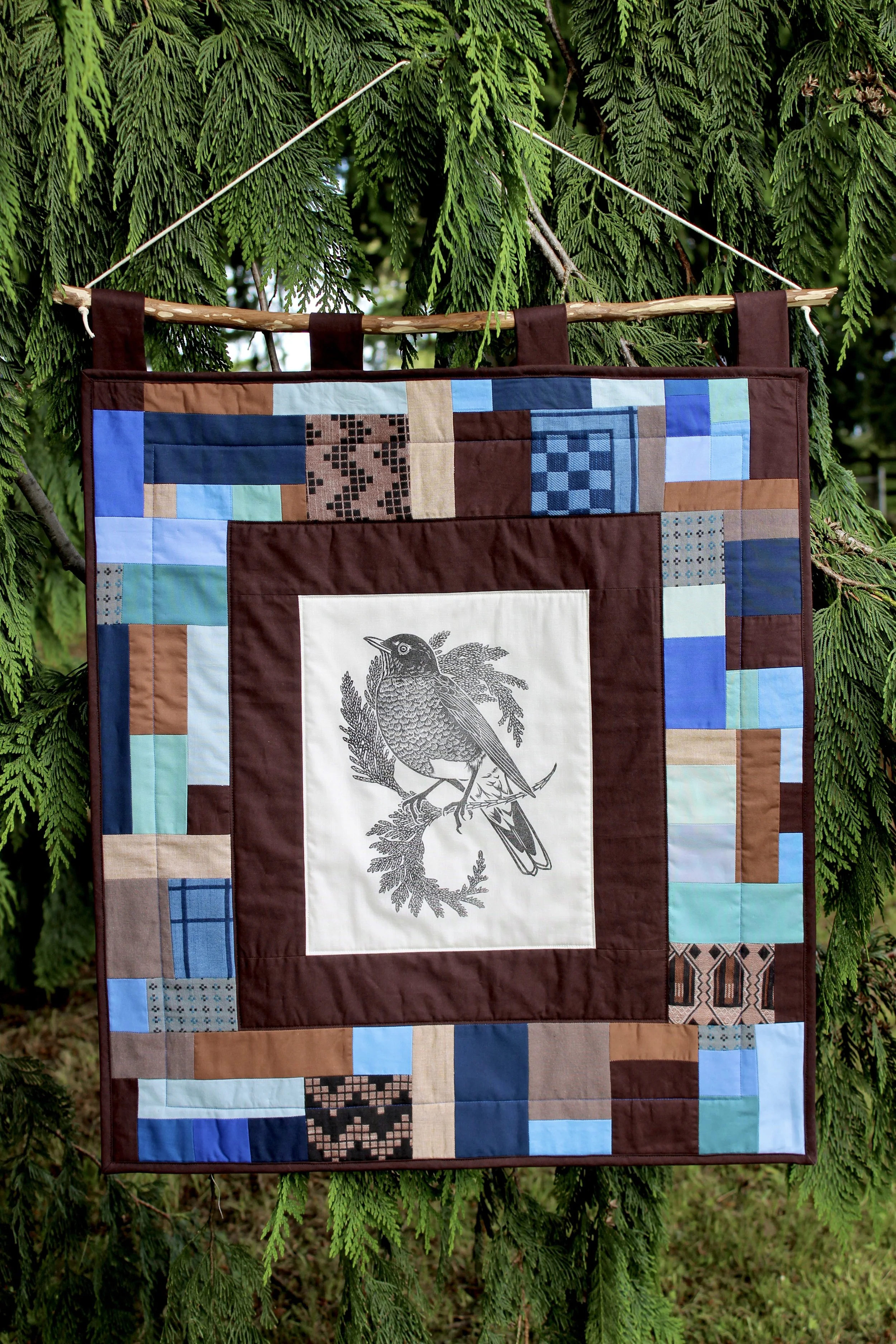 A quilt with a bird illustration hanging on a green tree.
