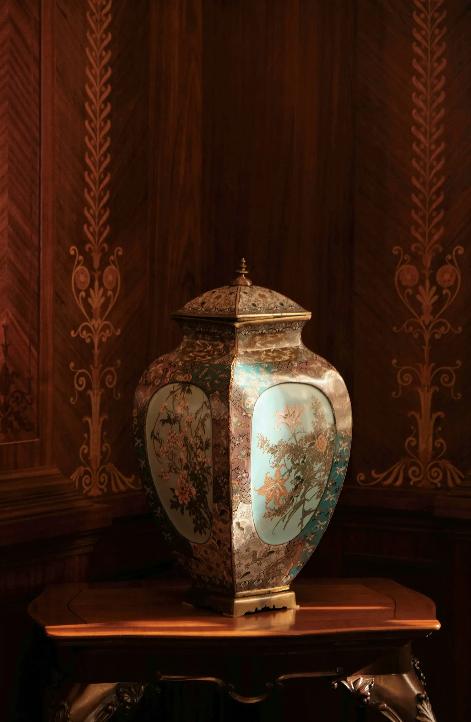 An ornate, painted porcelain lamp with a floral and landscape design, placed on a wooden surface against a dark, richly decorated wood-paneled wall.