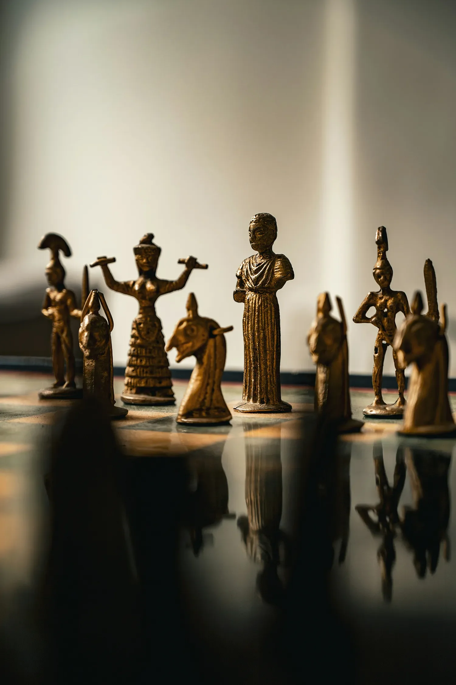 A close-up of a chessboard with ornate metal chess pieces including a king, queens, knights, and pawns. The pieces are reflected on the surface of the board, and the background is blurred with a neutral gradient.