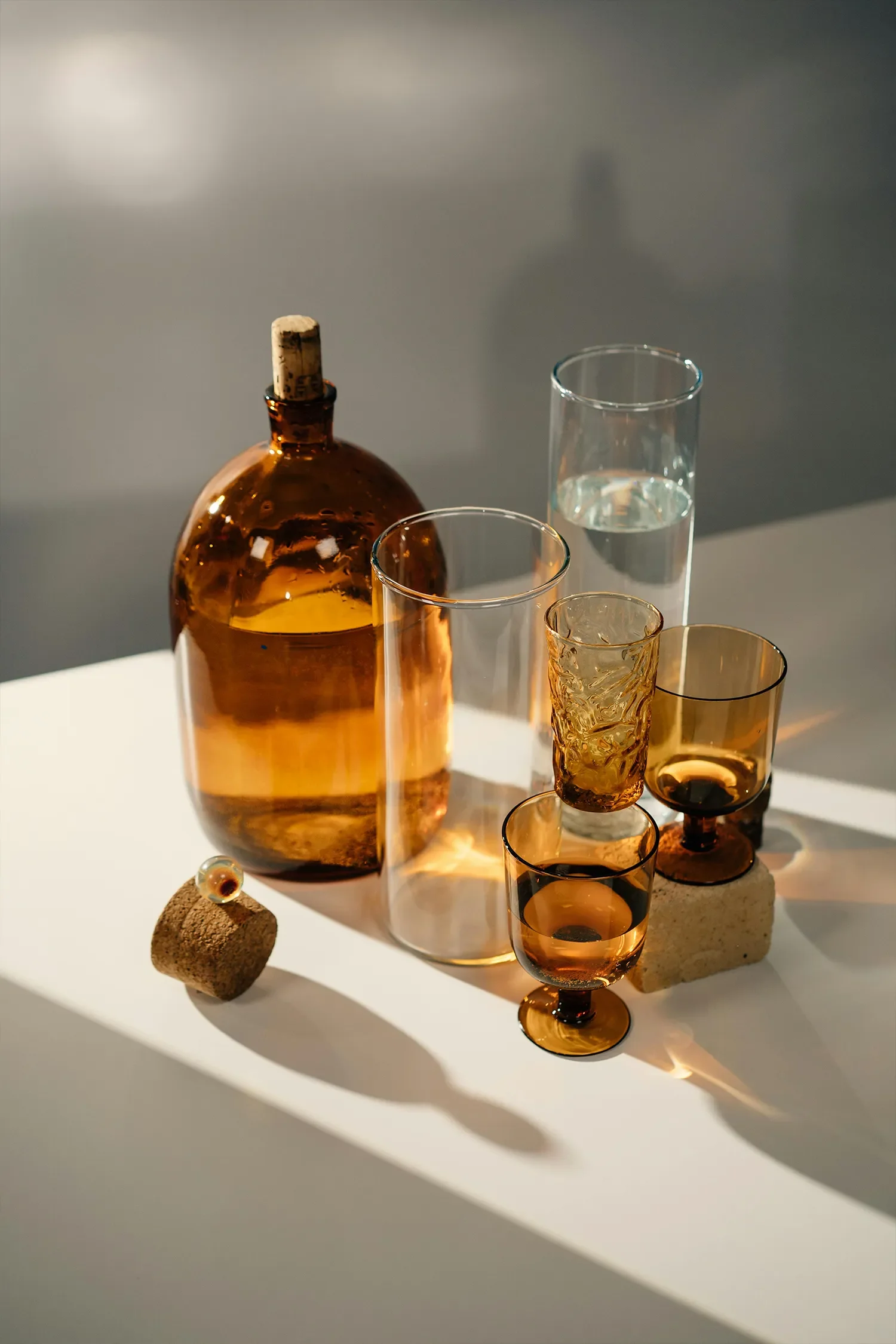 A collection of glassware, including a brown glass bottle with a cork, a tall clear glass containing water, two amber-colored glasses, and a textured amber glass, arranged on a white surface. There is a small cork stopper and a glass marble in the foreground. Shadows cast on a light gray background.