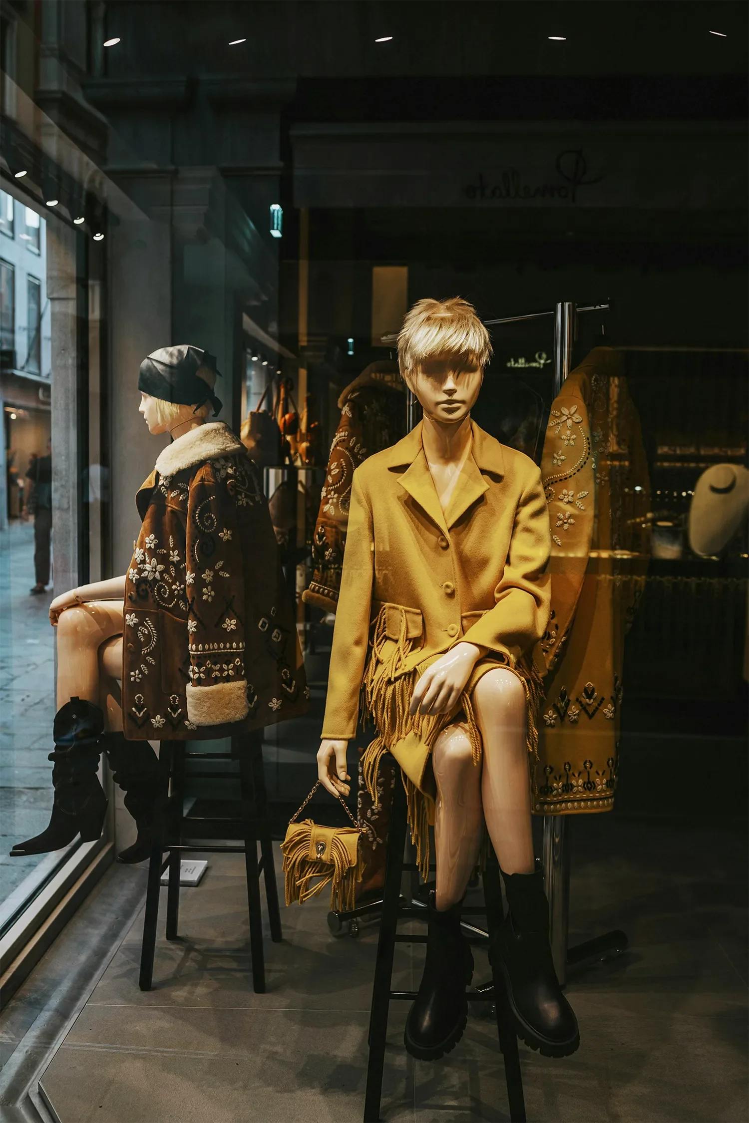 Display window with mannequins dressed in winter clothing, including a mustard coat and a fringed handbag, in an urban shopping district.