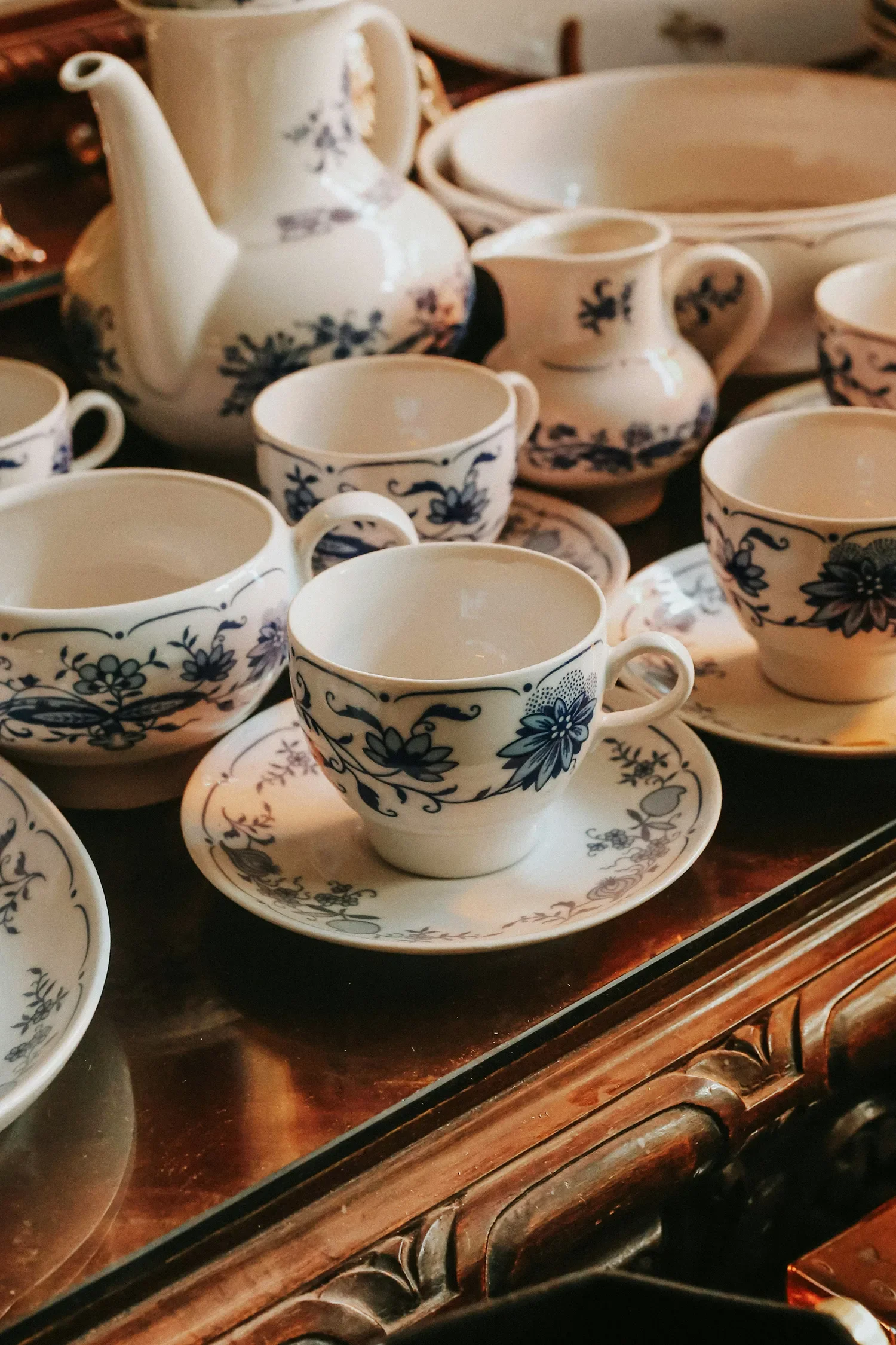 A collection of white porcelain teacups, saucers, a sugar bowl, a small pitcher, and a teapot with blue floral patterns arranged on a wooden surface.