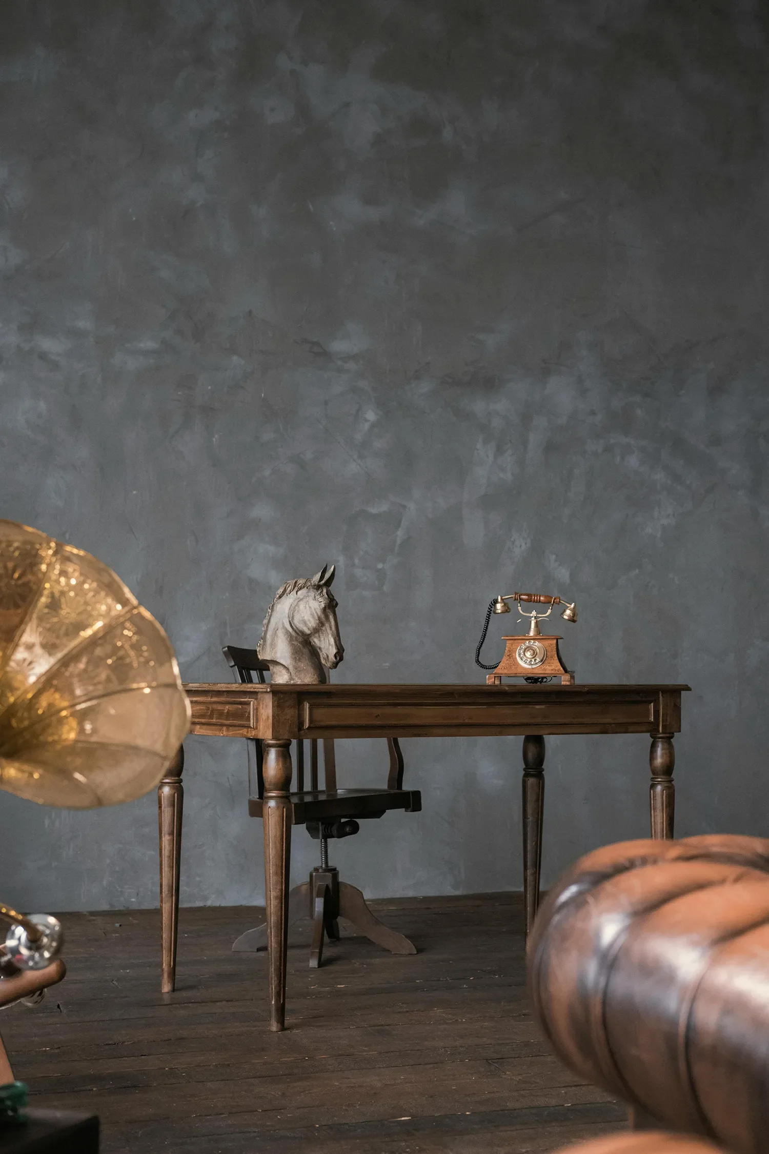 A room with a dark blue/gray textured wall, a wooden table, a vintage rotary telephone, a horse head sculpture, a black chair, and parts of brass table lamps.