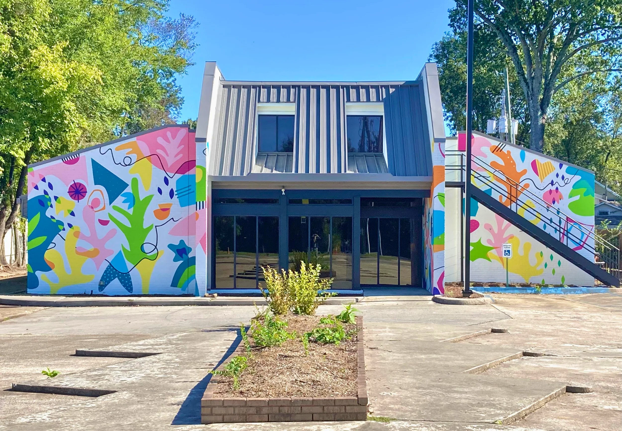 A modern building with a colorful abstract mural on the sides, metal roof with two large windows, and a small parking lot in front, surrounded by trees and greenery on a bright sunny day.
