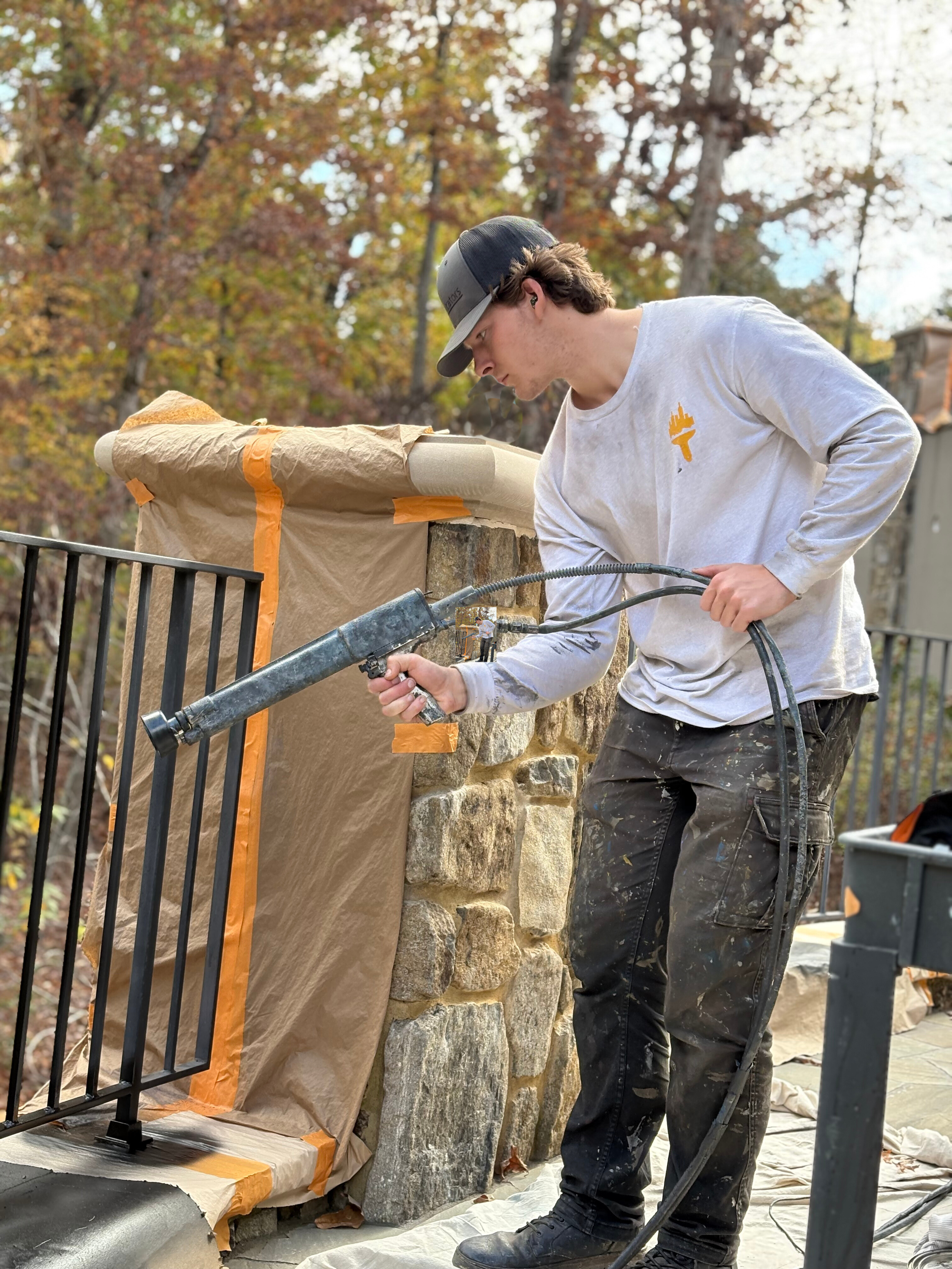 A young man in a gray shirt and black pants is applying sealant or filler to a stone wall with a power tool. He is outdoors with trees showing autumn foliage in the background.