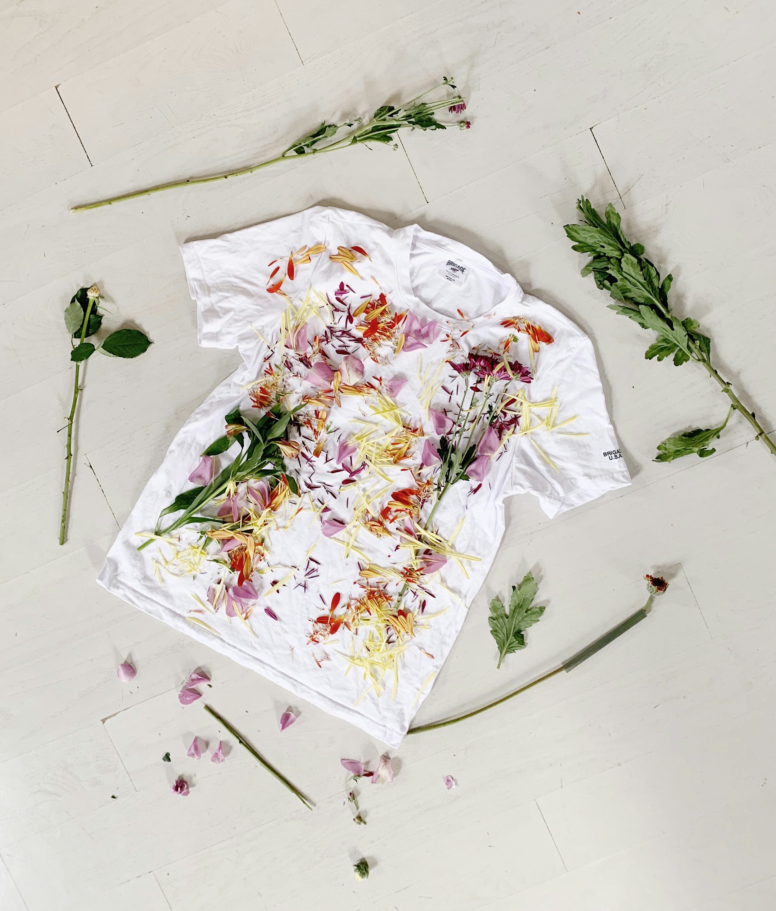 A white T-shirt covered with various flower petals and surrounded by loose flowers and green foliage on a light-colored wooden floor.