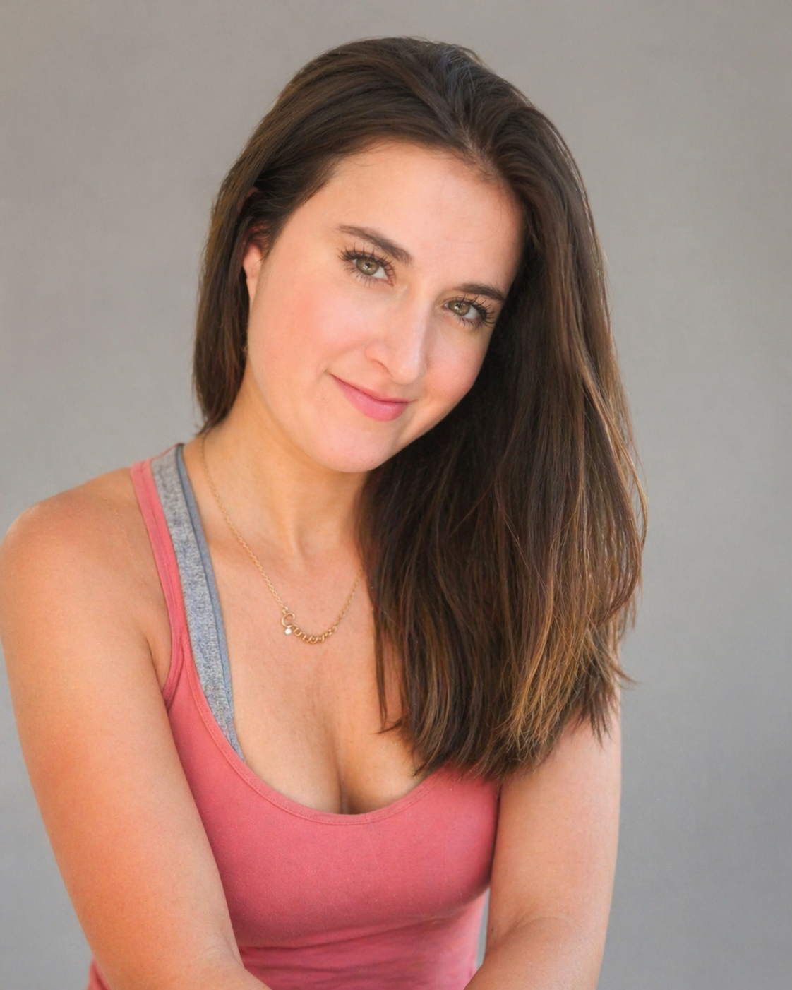 A woman with long brown hair, hazel eyes, and light makeup, wearing a pink tank top and a gold necklace, smiling and looking at the camera against a gray background.