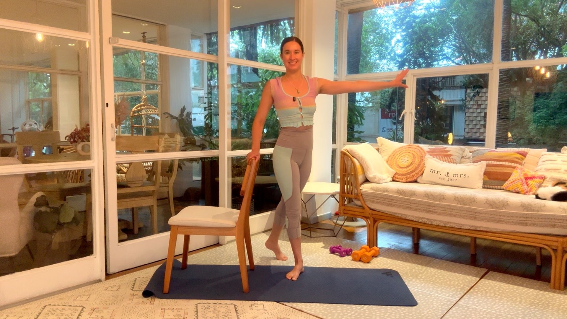 A woman is standing on a yoga mat in a sunlit room, holding onto a chair for balance, with workout dumbbells on the floor nearby. The room has large windows showing greenery outside, and a sofa with colorful pillows including one that reads ‘mr. & mrs.’