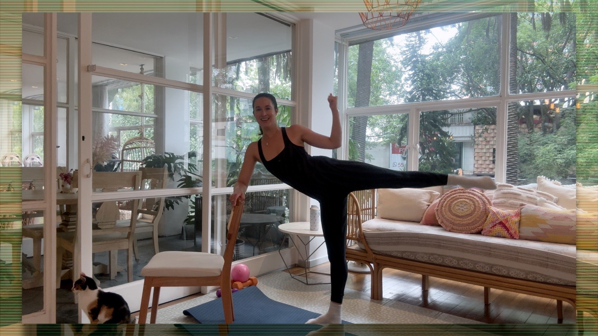 A woman practicing yoga indoors, standing on one leg with the other leg extended horizontally, holding onto a chair for balance. She is smiling, wearing a black tank top and black leggings. The room has large windows with a view of trees outside, and a cat sitting on the floor near a chair.