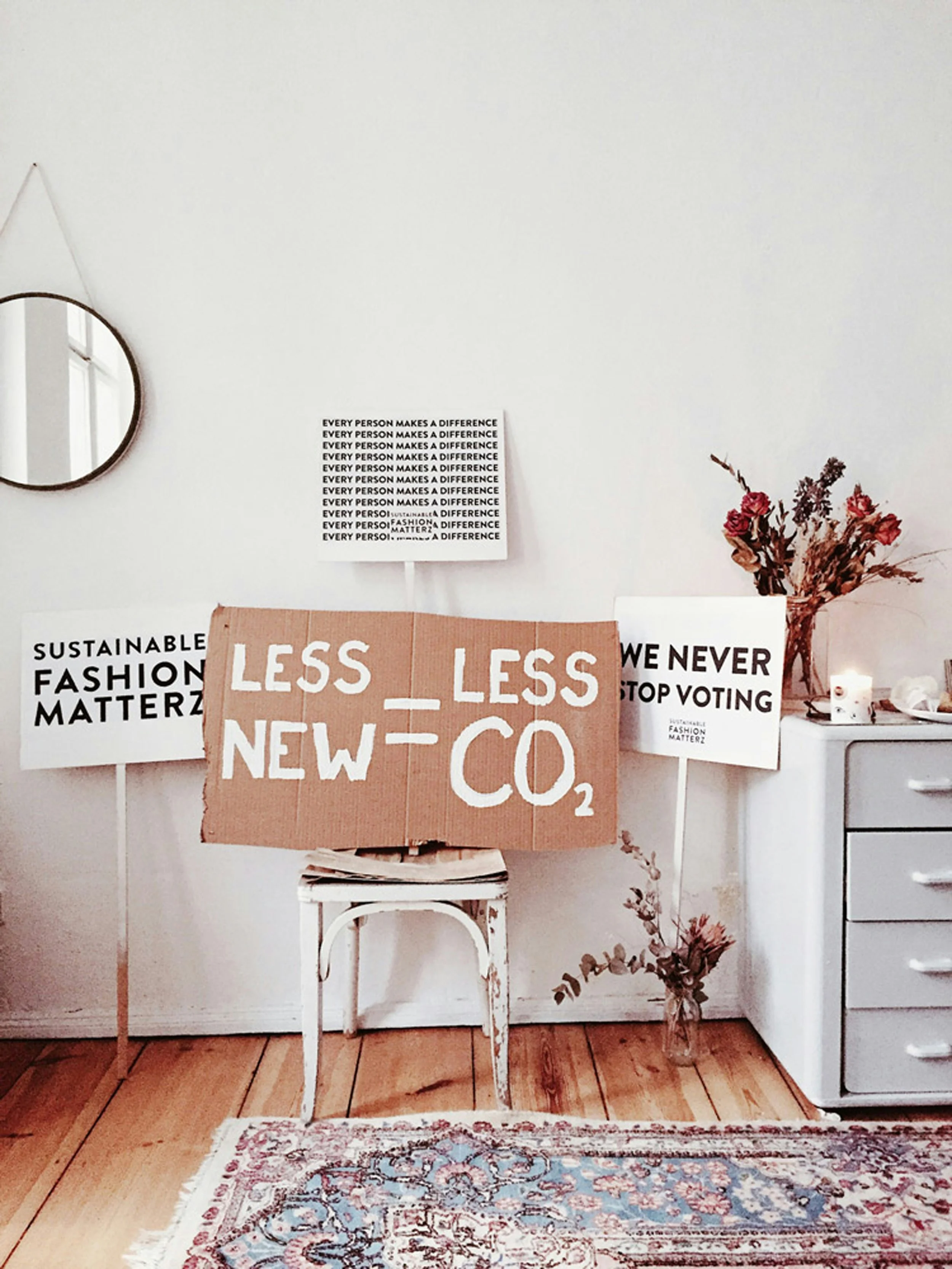 A room with protest signs advocating for sustainability and political change. Signs include messages about sustainable fashion, reducing carbon emissions, voting, and making a difference, with one featuring a cardboard sign stating 'Less Less New Co2'.
