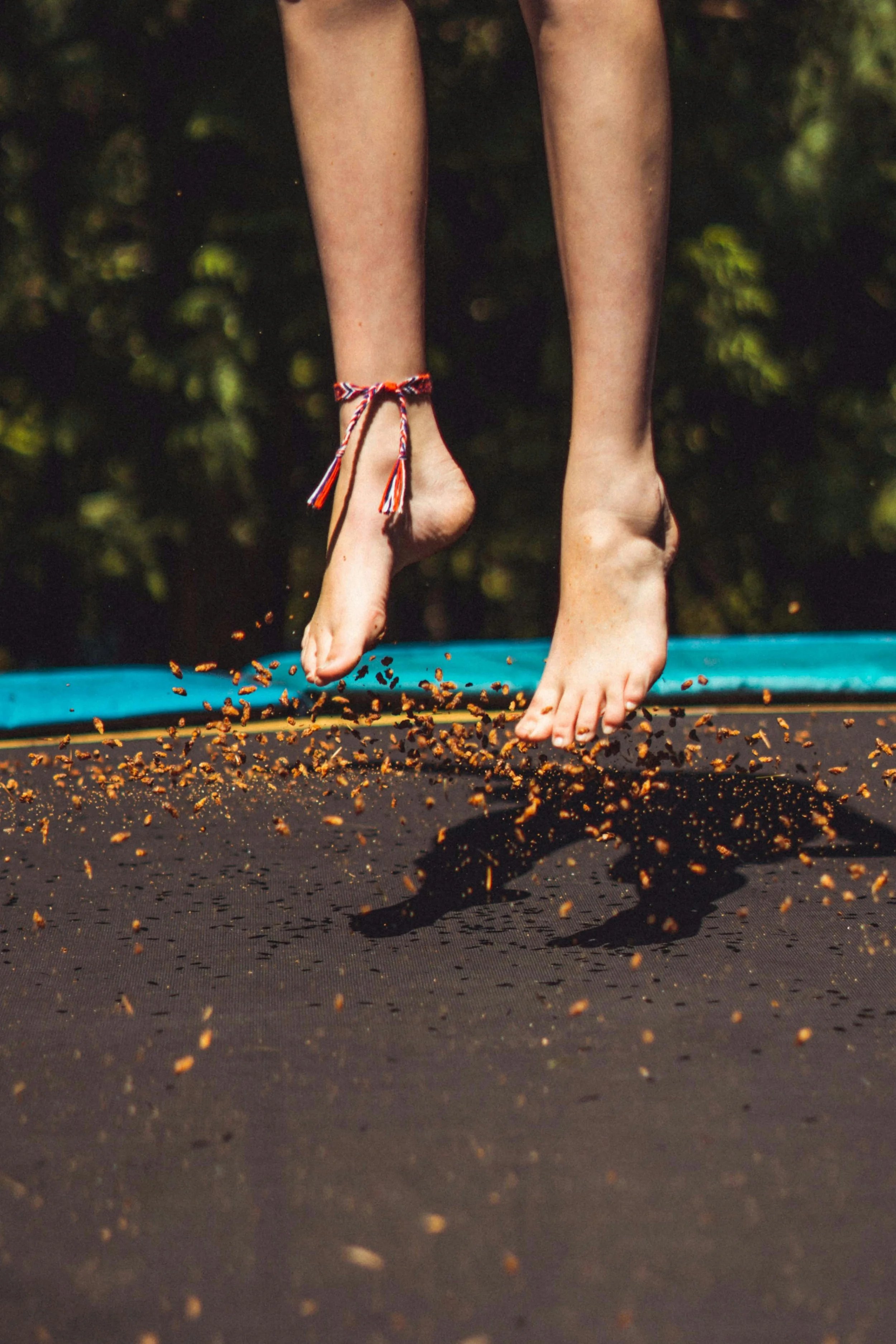 Person jumping on a trampoline, showing their legs and feet, with an ankle bracelet and dirt being kicked up from the trampoline surface.