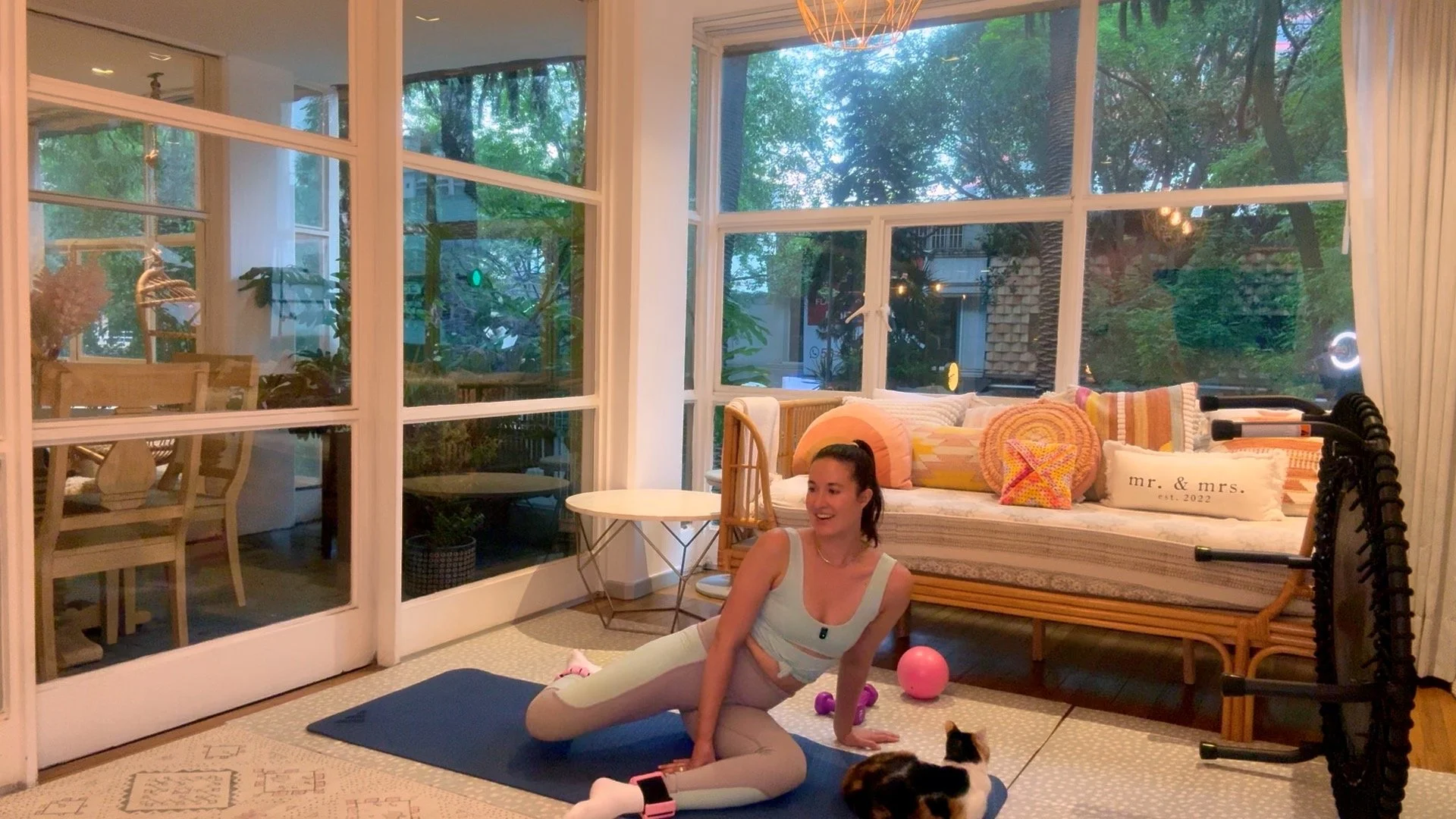 A woman practicing yoga on a black mat indoors in a sunny living room with large windows and green trees outside. She is wearing a light-colored sports top and leggings, smiling, with pink and purple exercise balls nearby. The room has a sofa with colorful pillows, a white side table, and a decorative light fixture on the ceiling.