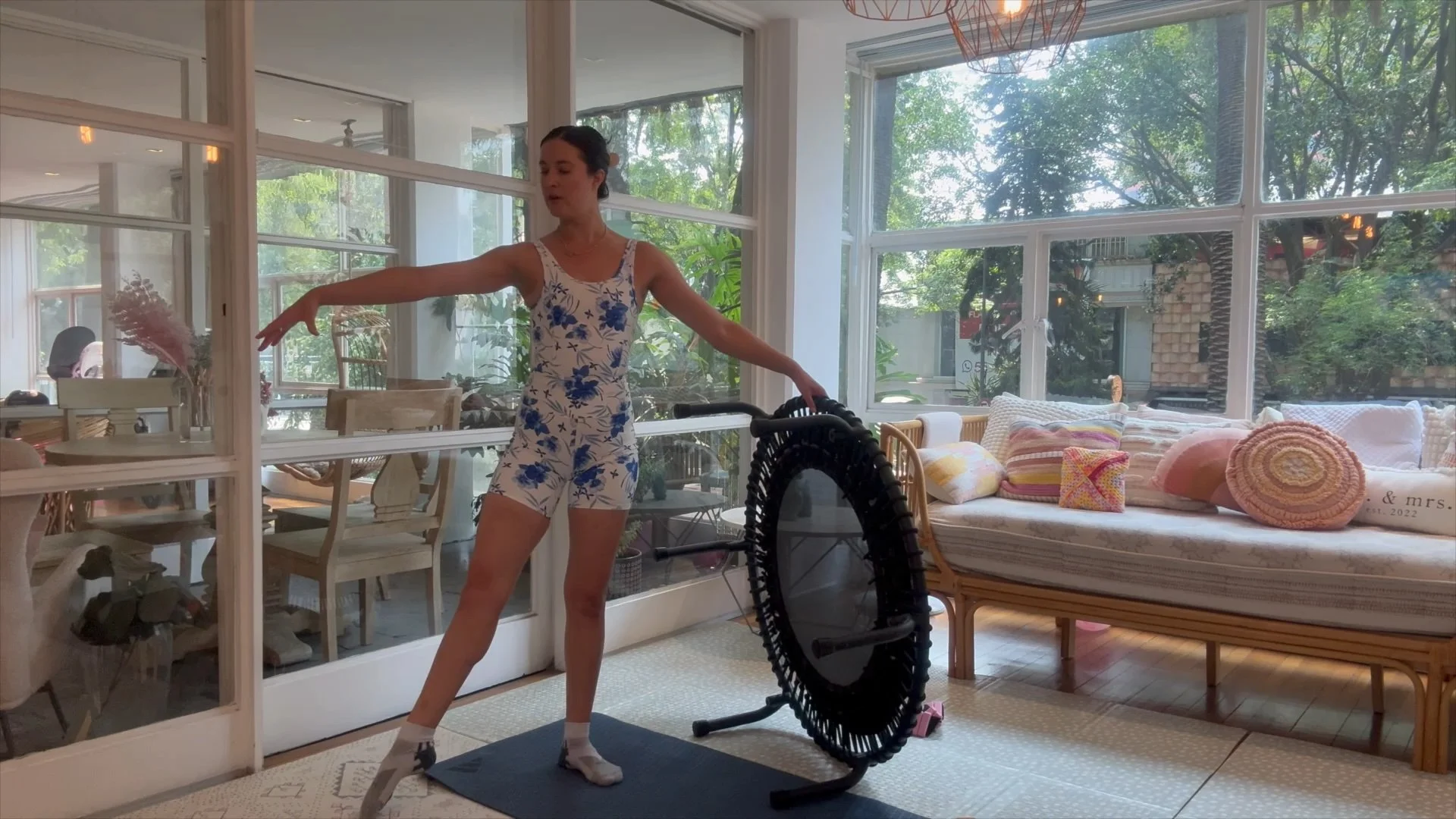 A woman in a floral sports outfit stretching in a bright, sunlit indoor space with large windows, a cozy sofa with colorful pillows, and a black exercise hoop in front of her.
