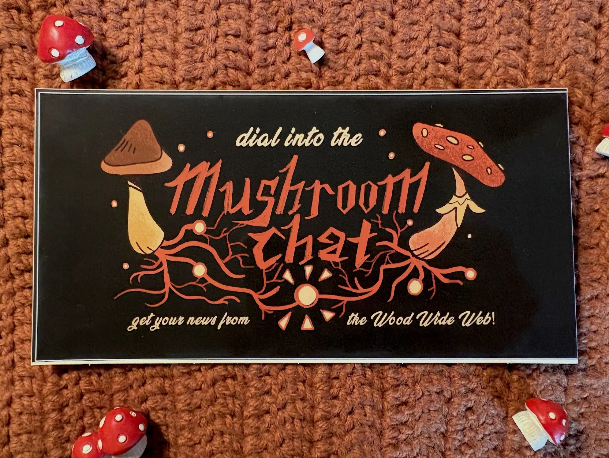 A printed advertisement featuring a black background with orange and red mushroom illustrations and text that encourages viewers to 'dial into the Mushrooth Web' and 'get your news from the Wood Wide Web.' The ad is placed on a textured, reddish-brow