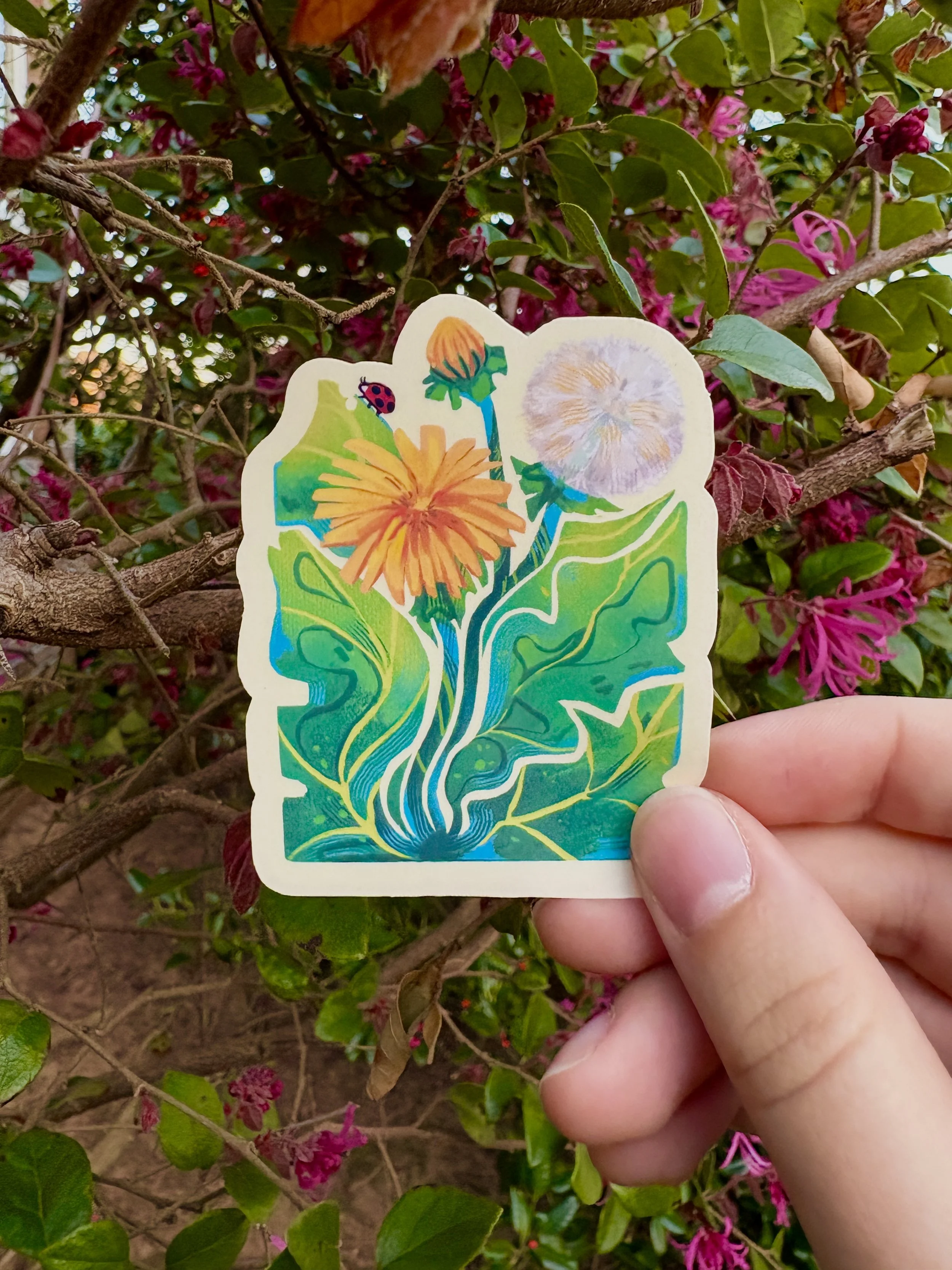 Colorful sticker of a dandelion and flowers, held in front of a leafy, flowering shrub with pink blossoms.