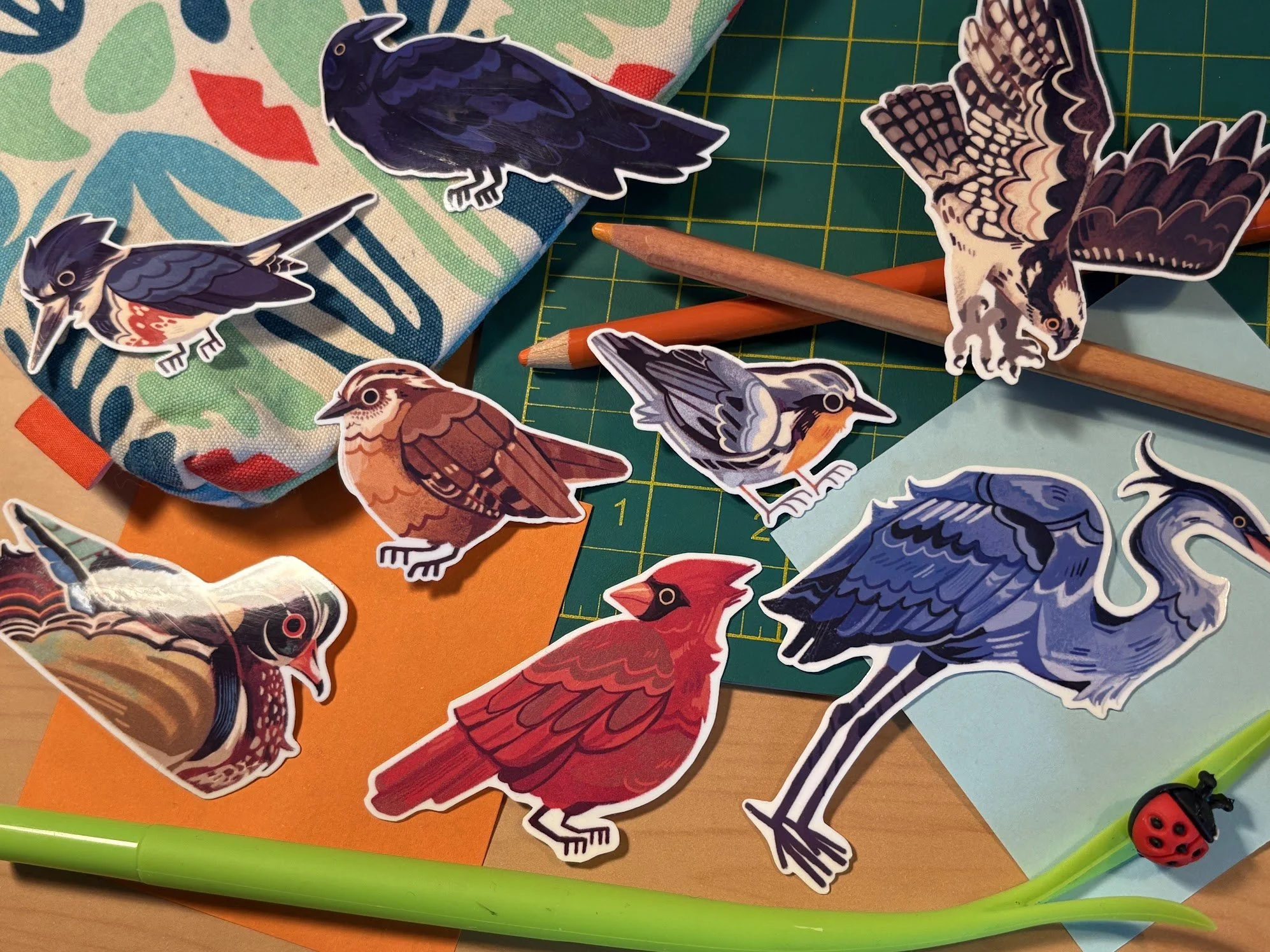 Assorted paper stickers of various bird illustrations, laid out among colorful papers, pencils, and craft supplies on a workspace.