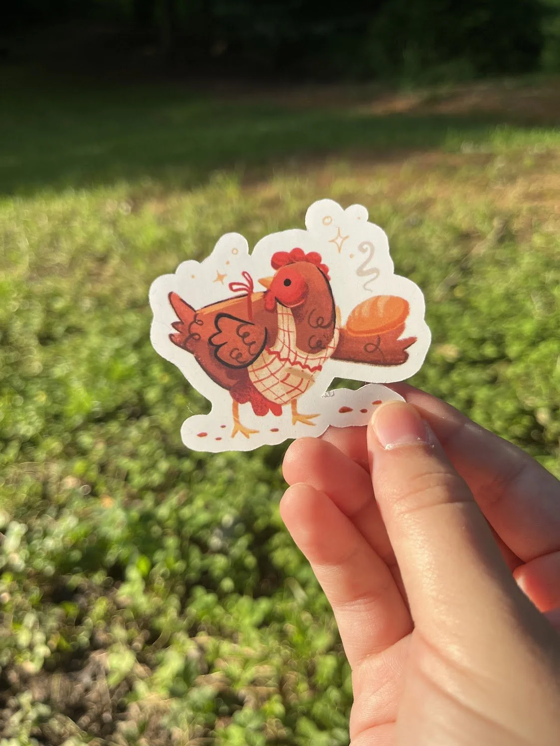 A paper cutout of a cartoon chicken held in a person's hand outdoors with green grass and trees in the background.