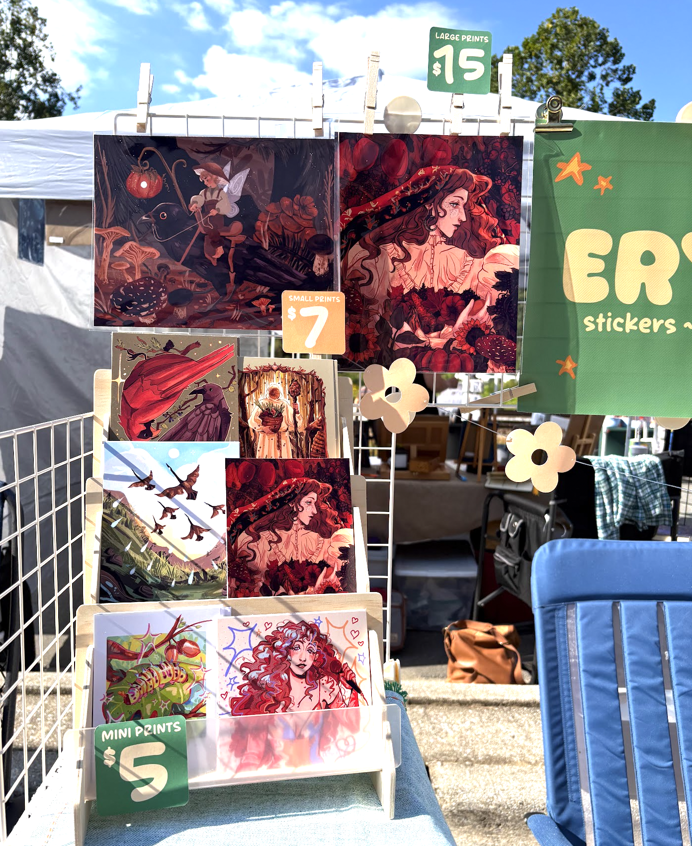 At an outdoor craft fair, there are artwork prints on display on a small stand with a sign saying 'Small Prints $7' and larger prints with a sign saying 'Large Prints $15.' The prints feature illustrations of fantasy characters and nature scenes in vivid colors. A blue chair is in the foreground, and there are other stall tents and trees in the background under a sunny sky.