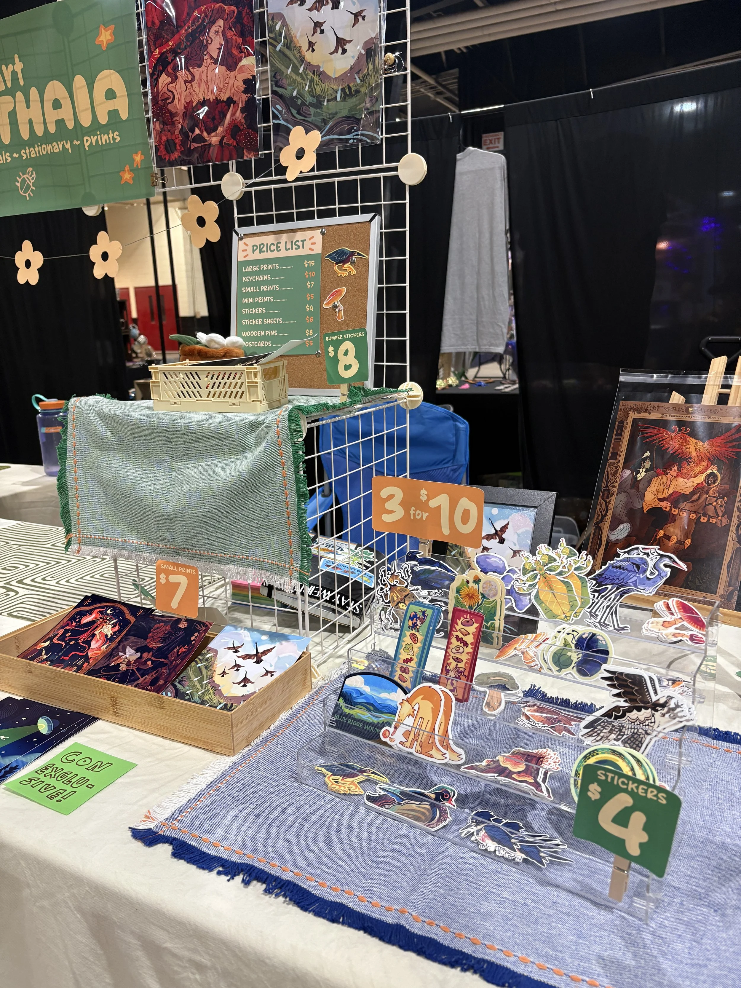 A display table at a craft fair with various stickers, prints, and pins featuring colorful fantasy and nature designs. There are price signs for items, a corkboard with a price list, and hanging artwork in the background.