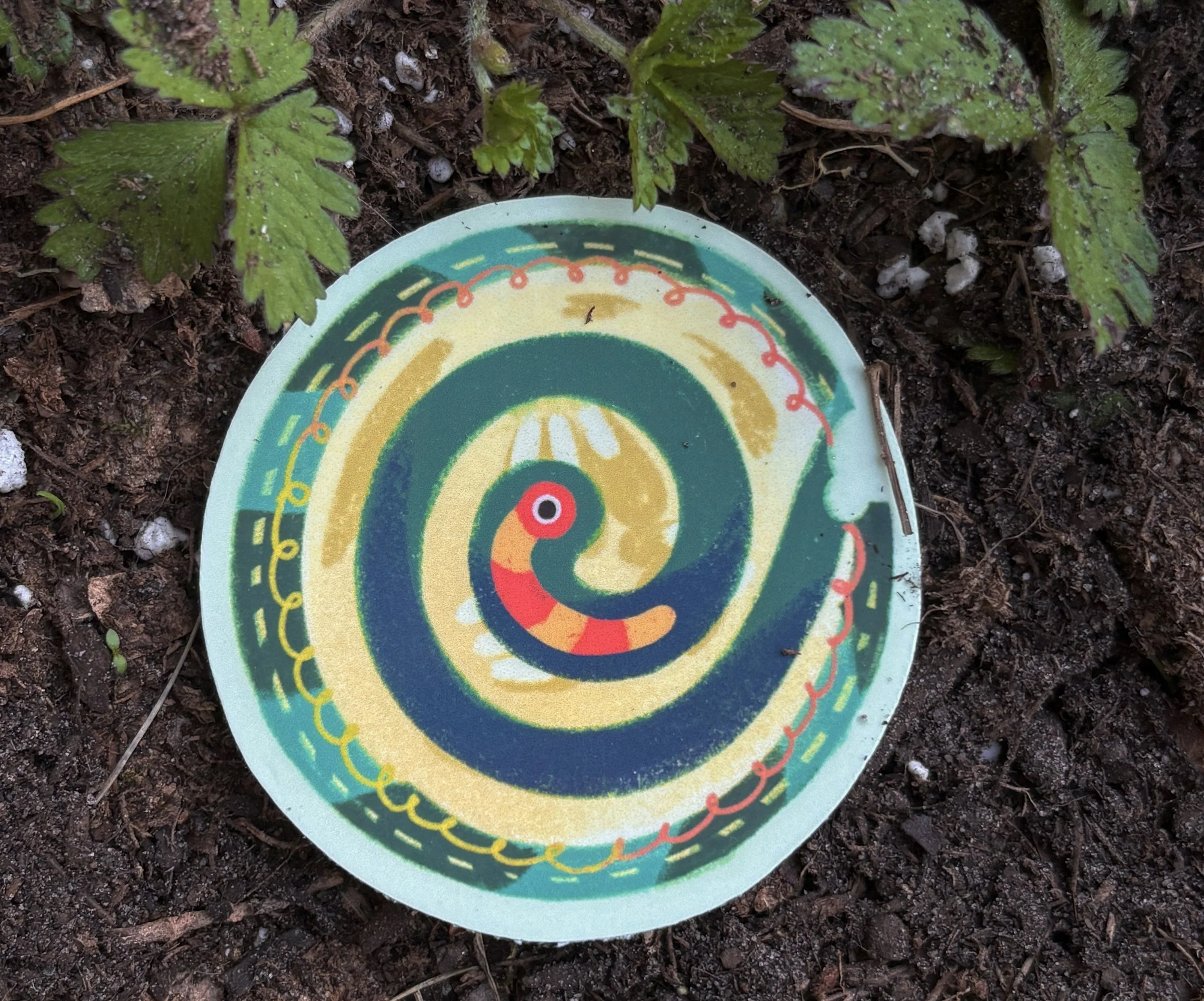 Colorful weather vane on the ground surrounded by soil and small green plants.