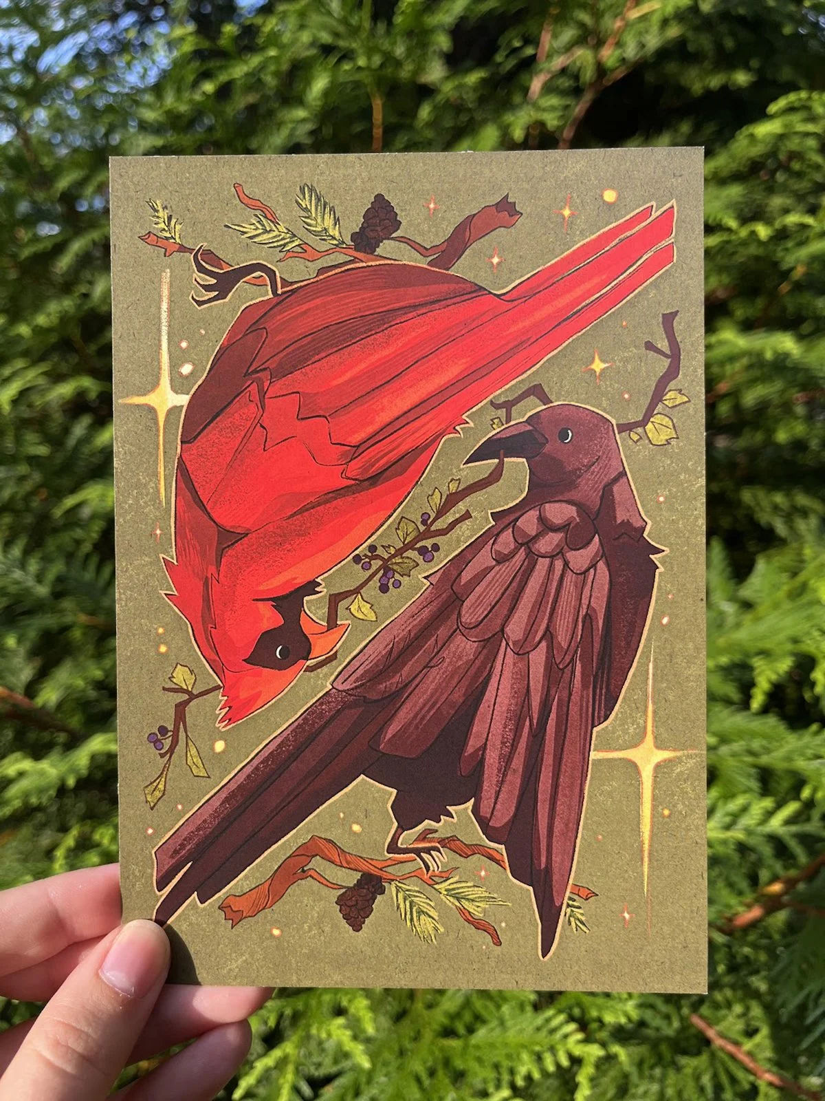A hand holding an illustrated card featuring a red bird and a brown bird with a background of green foliage. The illustration includes stars and decorative elements.