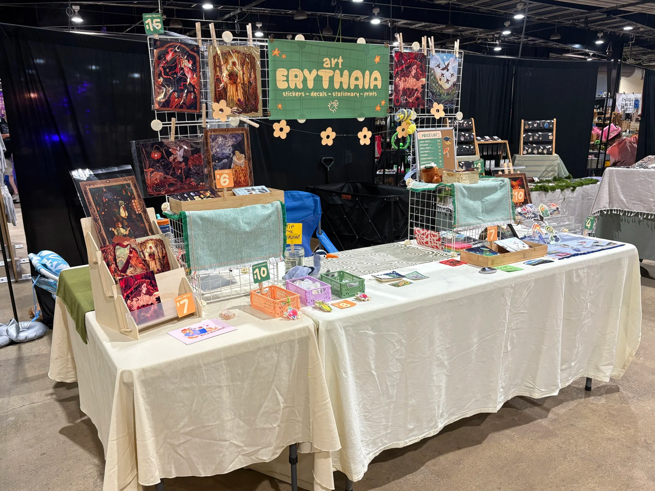 A booth at an art fair displaying various artworks, stickers, and stationary items, with signs indicating prices and a large green banner reading 'Art Erythnia'.