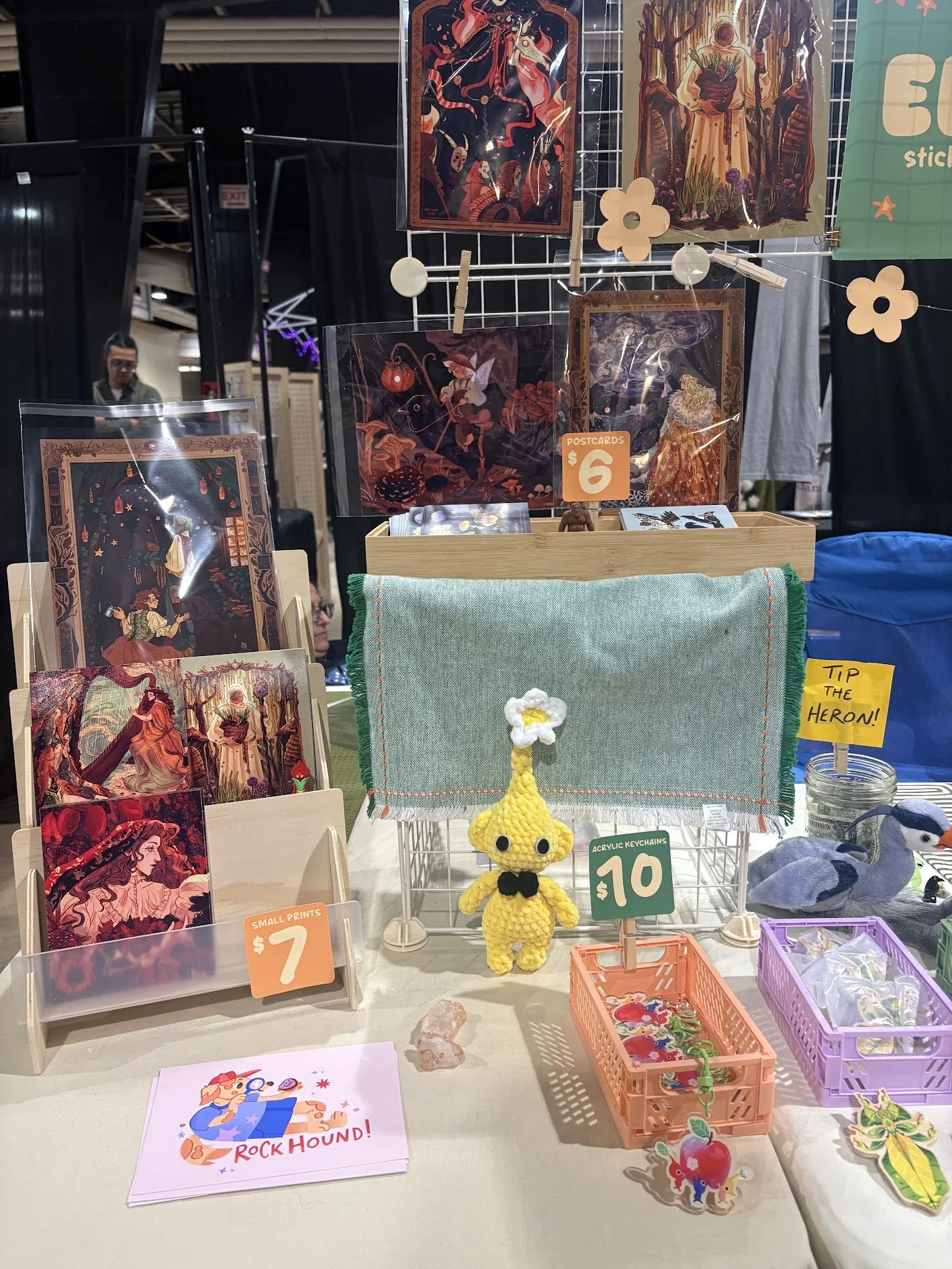 A display booth with fantasy-themed posters, artwork, and handmade items including a yellow crocheted duck keychain, small prints, stickers, and a fabric piece with a flower embellishment. Price tags indicate items are sold for $6, $7, $10, and $12.