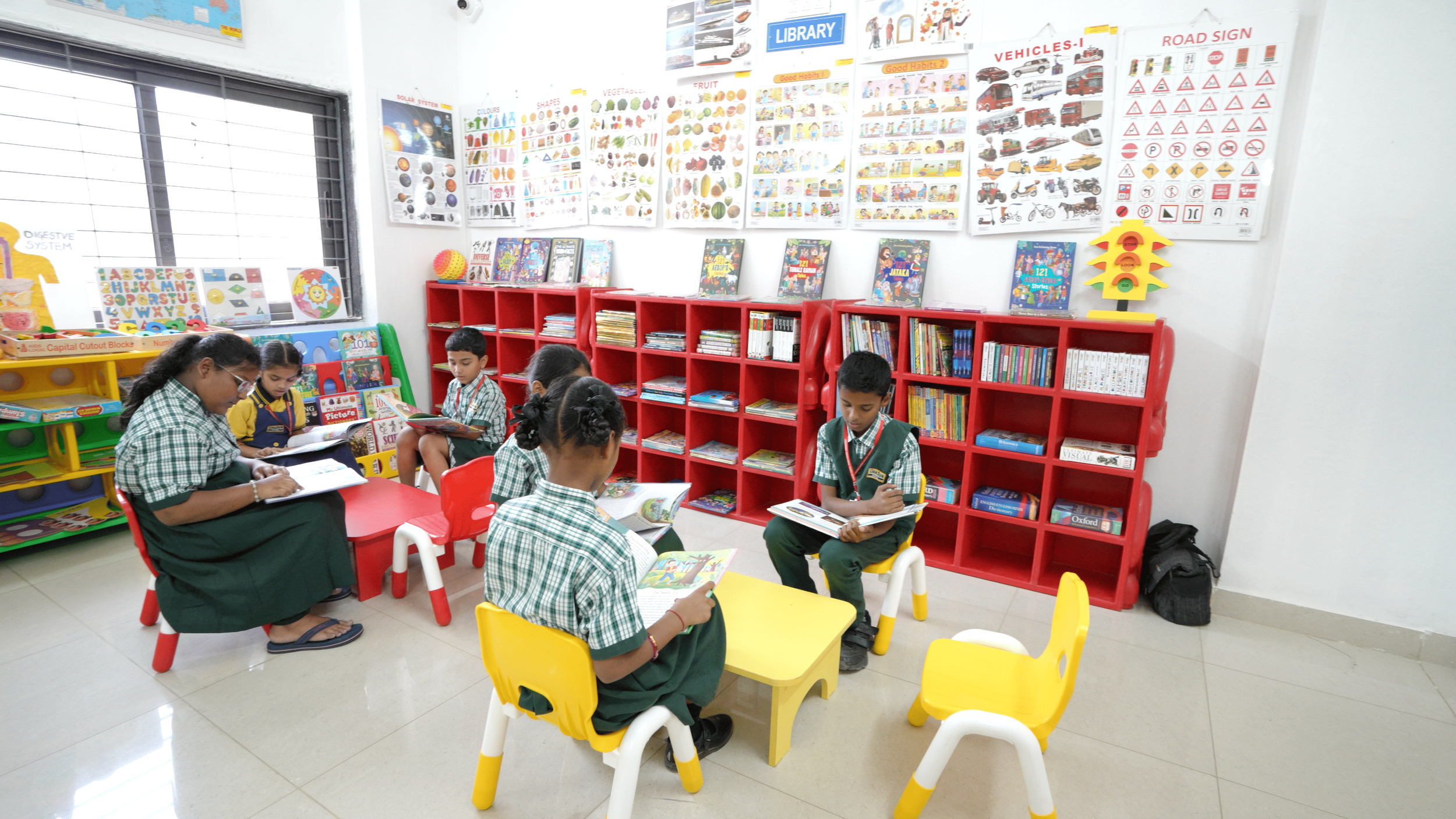 Children in school uniforms reading books in a library classroom with red bookshelves and educational posters on the wall.