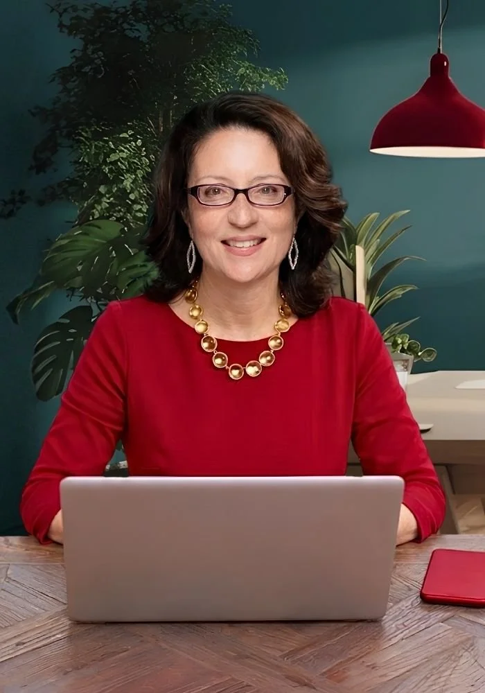 Wendy Maldonado sitting at a wooden desk in front of a silver laptop, wearing a red top, a gold beaded necklace, and large earrings, in a room with dark green walls, potted plants, and a red pendant light.