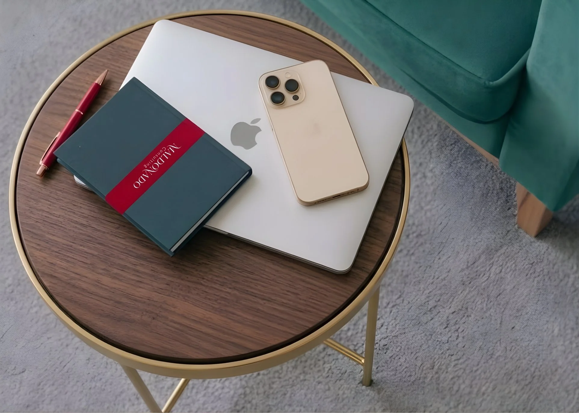 An oval wooden table with gold-colored legs holds a closed gray Vaionland notebook, a red pen, a white laptop with a silver Apple logo, and a  beige iPhone with three camera lenses. The table is situated on a light gray carpet near a teal upholstered chair.