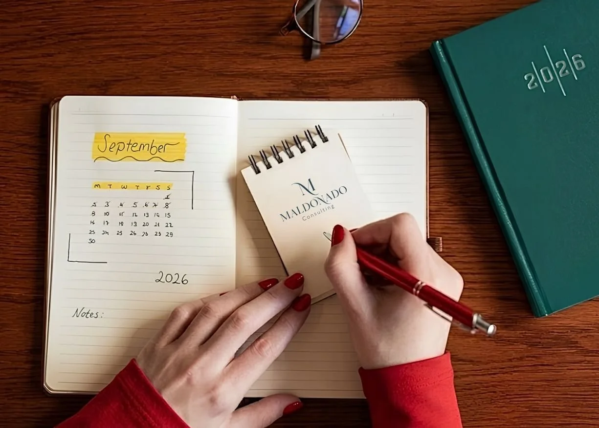 Open notebook with September 2026 calendar, notes section, a person's hands writing in a small notepad labeled "Maldonado Consulting" on a wooden desk with a teal book and glasses nearby.